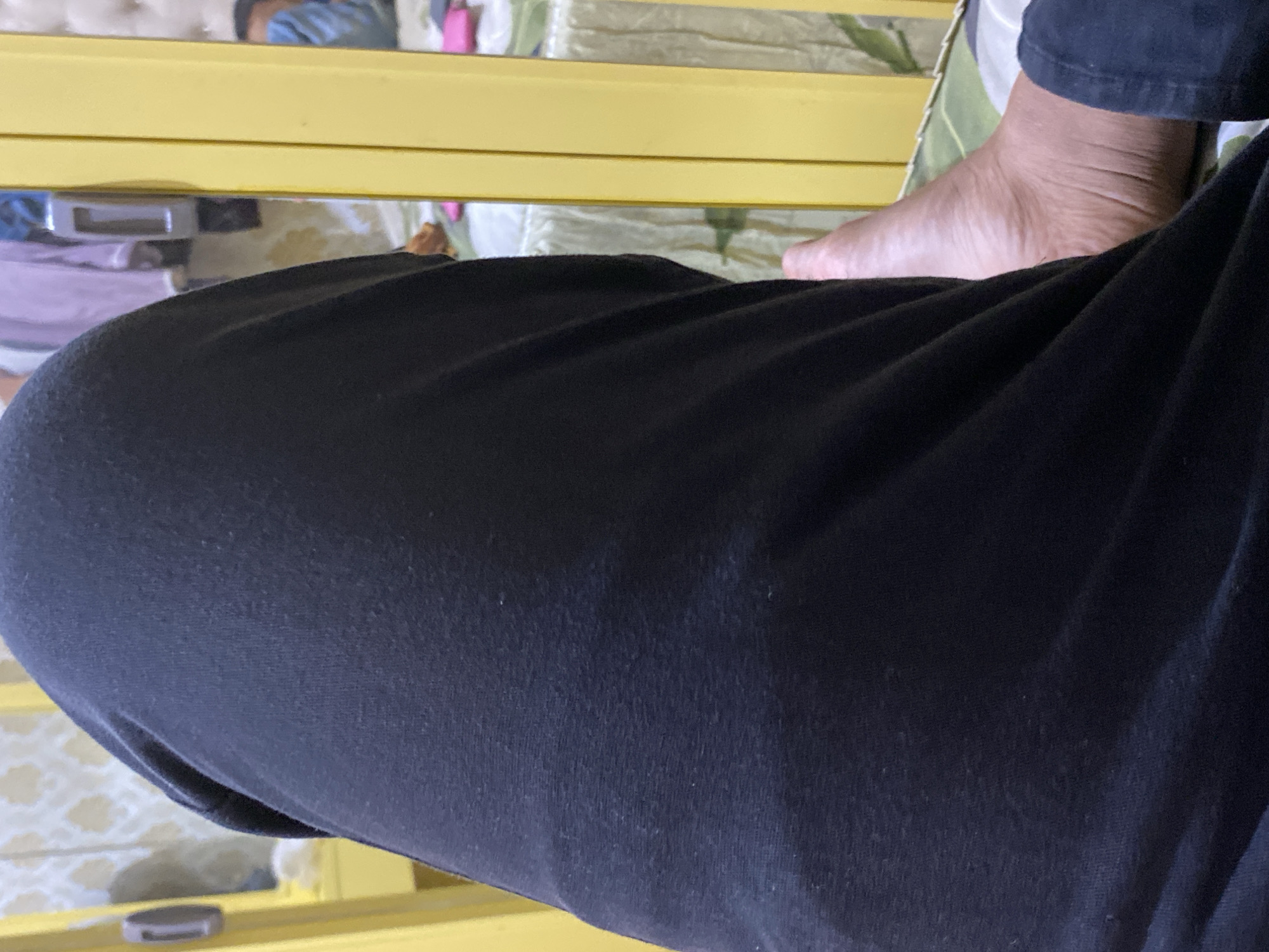 The image shows a close-up view of a person's legs, with one leg prominently in the foreground covered by black pants. A bare foot, with wrinkled skin, is visible on the right side of the frame, resting on a patterned surface. In the background, a yellow structure, possibly a bed frame or furniture, is visible. The environment appears to be indoors, with hints of bedding and reflections in what might be a mirror or glass surface. The overall lighting suggests an indoor setting, possibly in the evening or a dimly lit room.