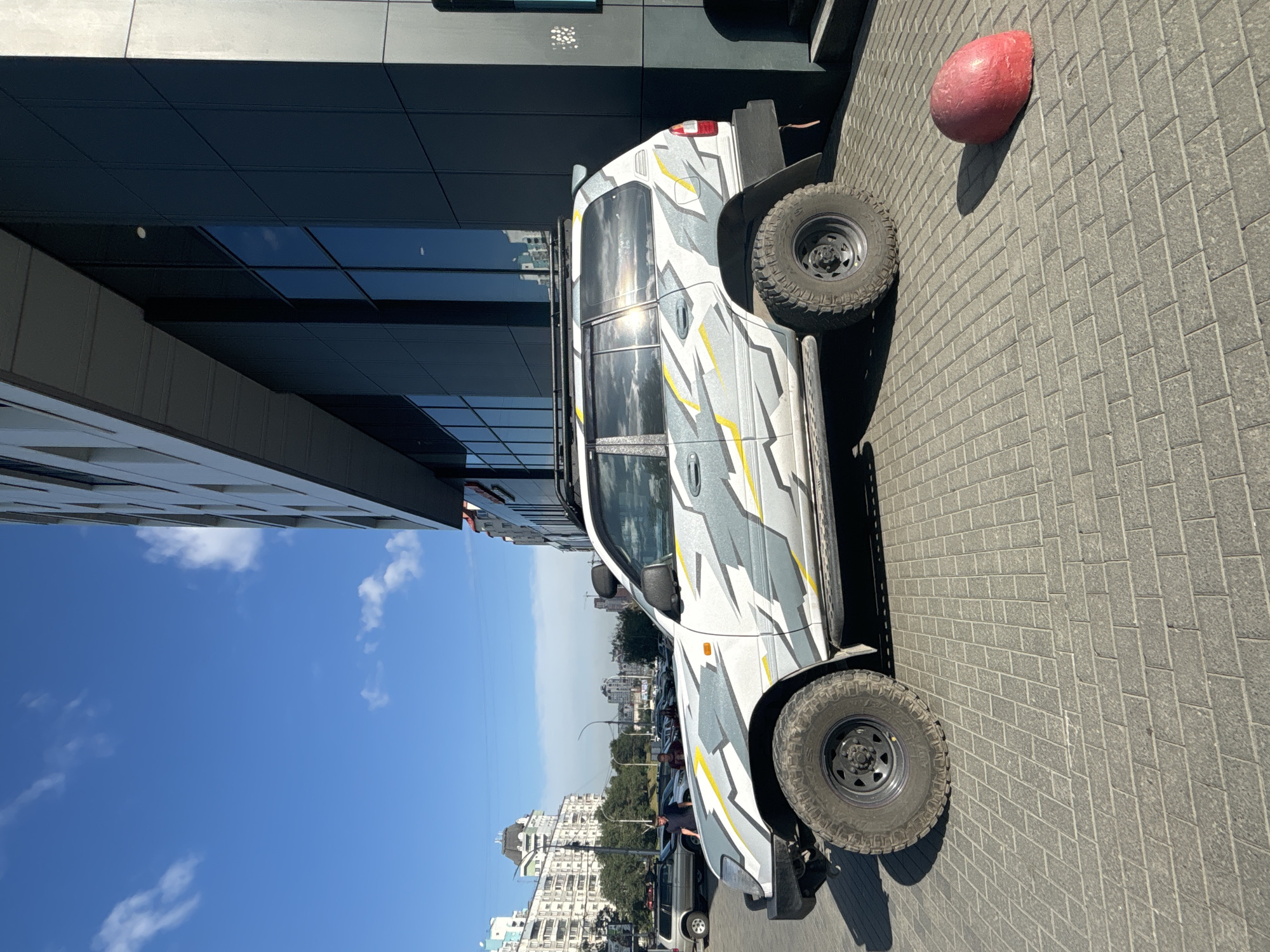 A white SUV with a gray and yellow camouflage wrap is parked on a paved sidewalk in front of a modern, dark-paneled building. The vehicle has large off-road tires and a roof rack. Sunlight glints off the windows of the SUV, reflecting the sky and clouds. To the right of the vehicle, a red, rounded bollard sits on the sidewalk. In the background, across a street, are several multi-story buildings, some appearing residential and others commercial, indicative of an urban environment. The sky is blue with scattered white clouds. The scene appears to be in the city of Novosibirsk, Russia, as indicated by the architecture and overall urban setting.