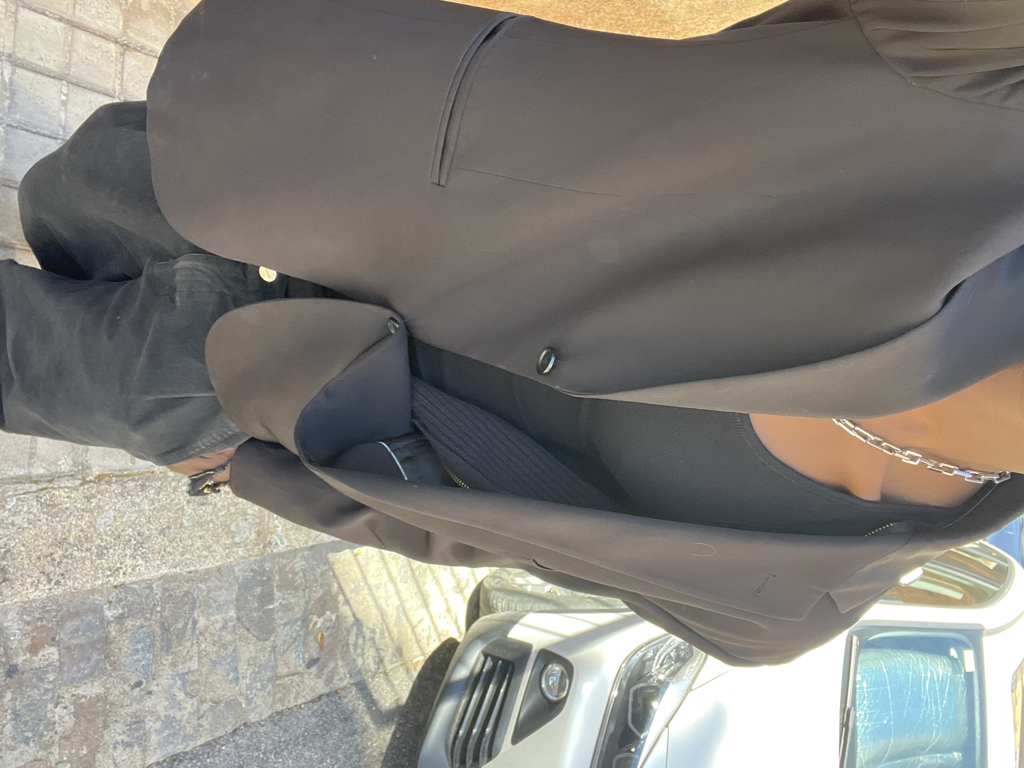 A medium, eye-level shot depicts a person standing on a pavement possibly in the city of Barcelona, Spain. The subject wears dark grey or black jeans and a matching blazer. The blazer is fastened by a single button. Beneath the blazer, a black garment with a ribbed texture is visible. The person also wears a silver chain necklace. In the background, a silver car is partially visible. The pavement consists of square grey paving stones.