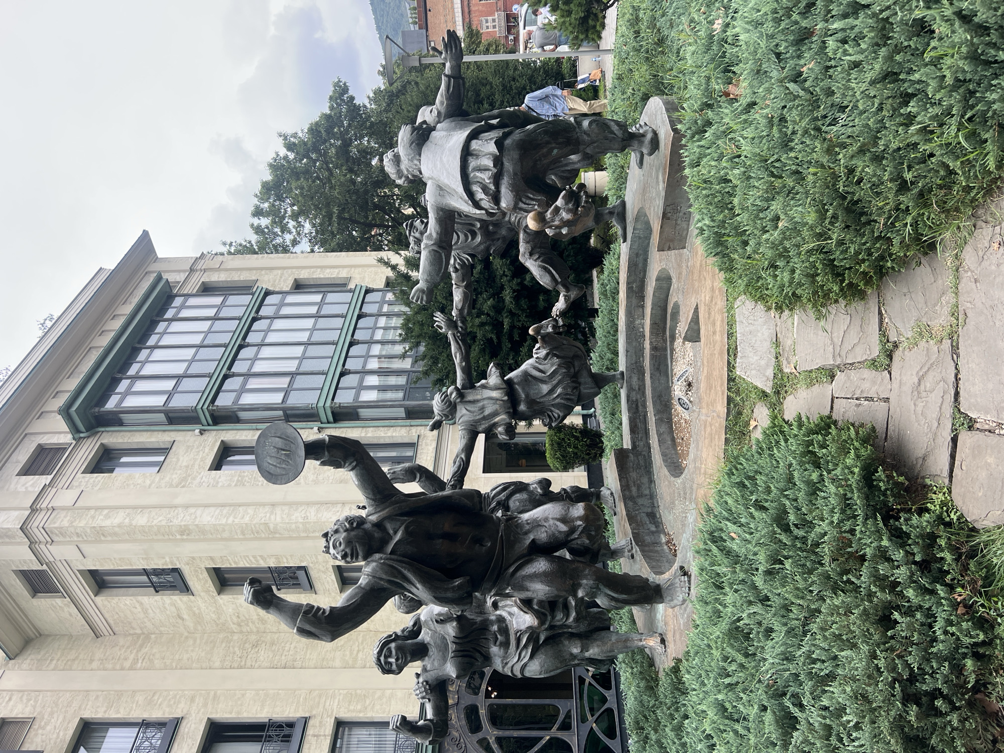 The image depicts a bronze sculpture of several figures dancing in a circle in front of a building. The sculpture is located in Tbilisi, Georgia, in front of the former Parliament building, now a hotel. The dancing figures appear to be in motion, with their arms outstretched and legs in various stages of movement. They are dressed in historical attire. The building in the background is multi-story with a light-colored facade and dark windows. The area around the sculpture is landscaped with lush green bushes and a stone pathway. The sky is overcast.