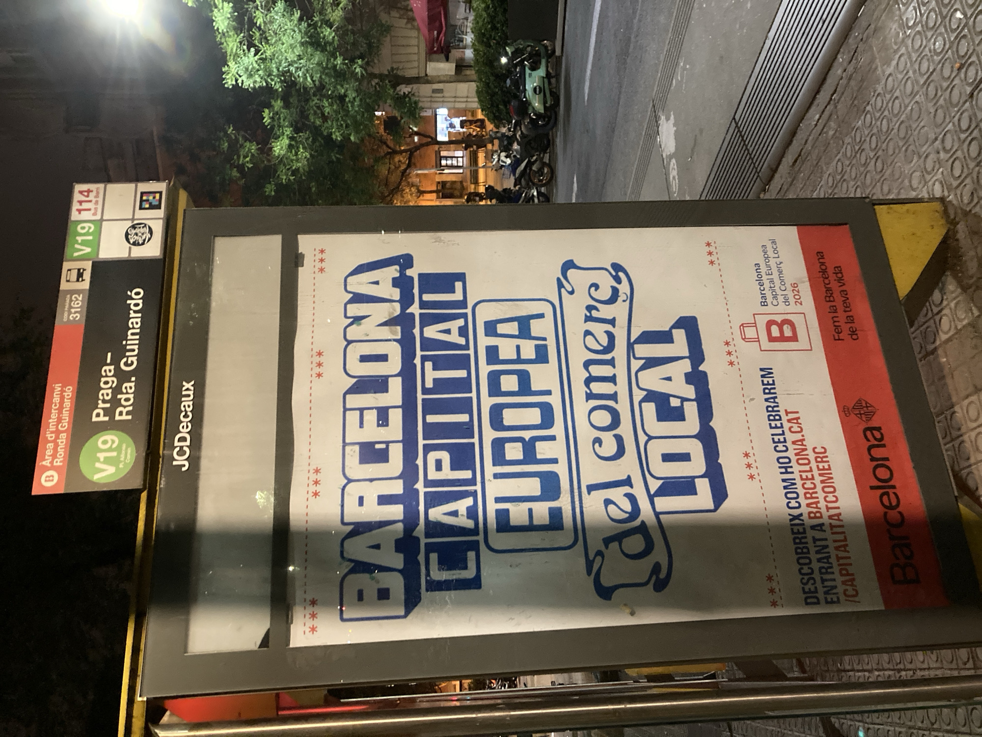 Here is a description of the image:
The image depicts a bus stop sign in Barcelona, Spain, with advertisements promoting the city's designation as the European Capital of Local Commerce in 2026. The sign lists bus routes V19 and 114, with "Praga - Ronda Guinardó" displayed as the bus stop name. The bus stop is also located near the "Area d'intercanvi Ronda Guinardó." A JCDecaux logo is visible on the sign. The advertisements on the sign emphasize "Barcelona Capital Europea del comerç Local." The image is taken at night, with street lights illuminating the scene. In the background, parked motorcycles and pedestrians are visible along the street.