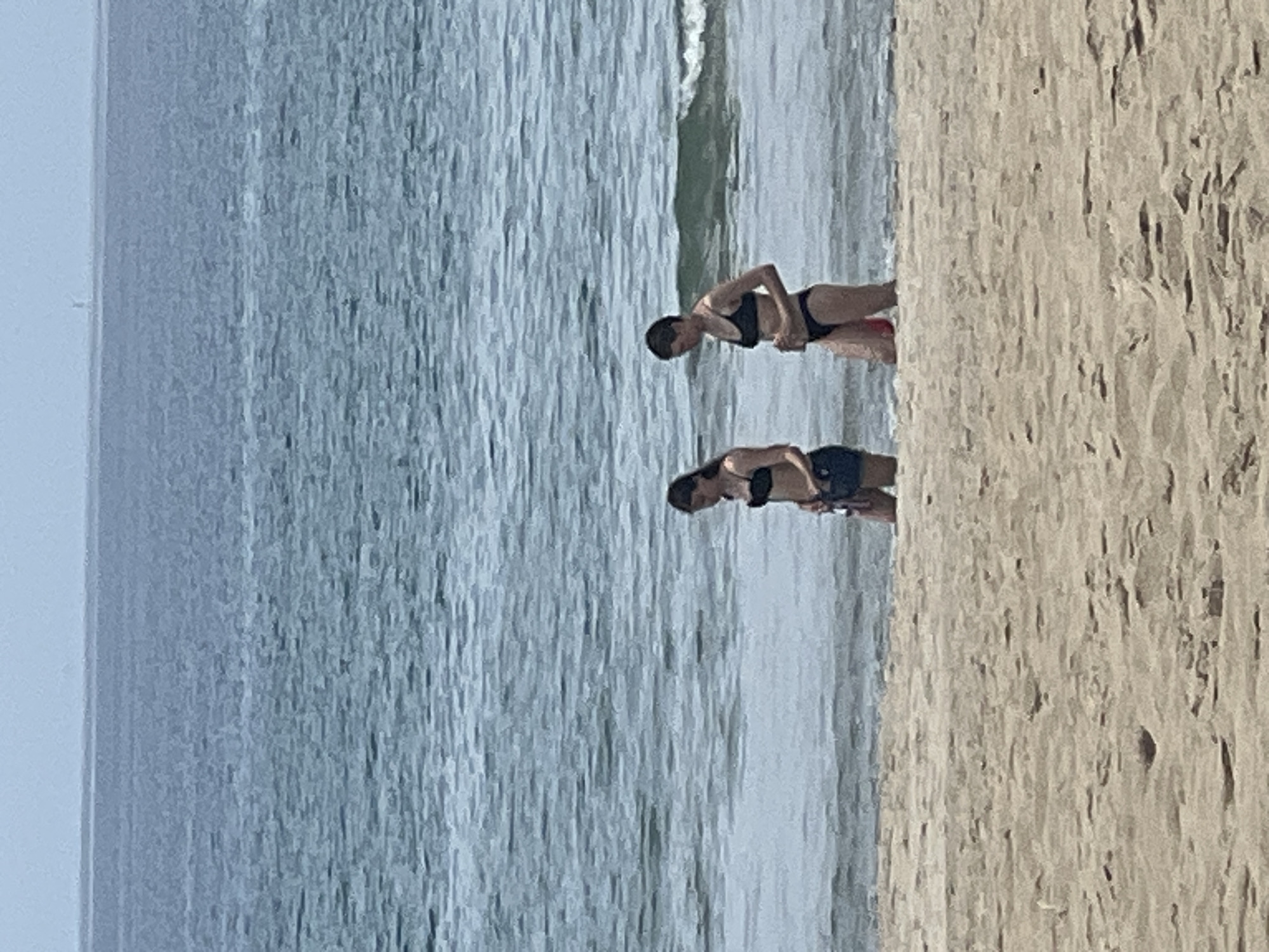Two people are wading in the shallow ocean water at My Khe Beach in Da Nang, Vietnam. The person on the left is wearing a black bikini and dark shorts, and the person on the right is wearing a black bikini. The sand in the foreground is light brown. The water is a blue-gray color, and there are small waves breaking. The horizon is visible in the distance.