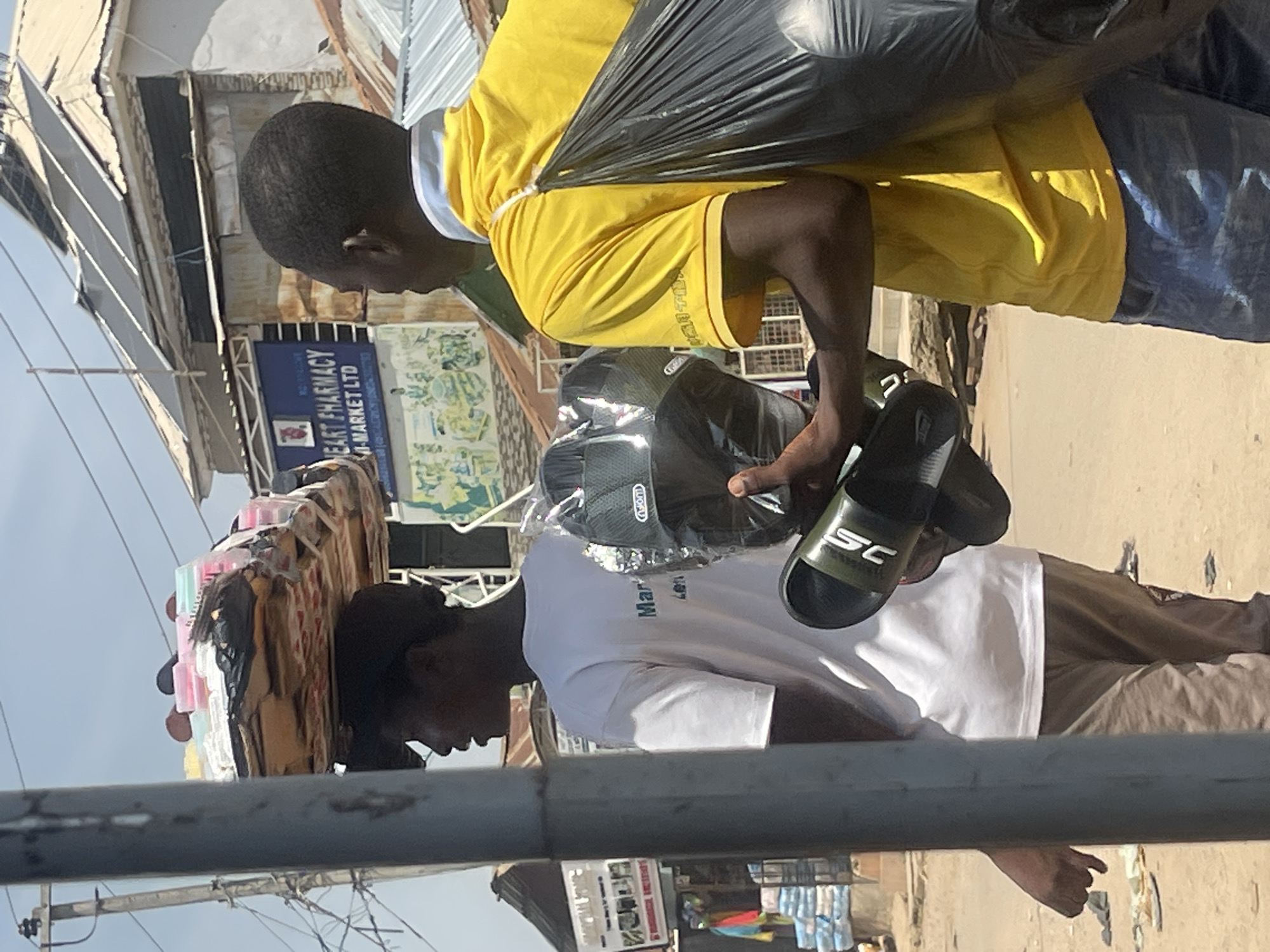 In Abuja, Nigeria, on a sunny day, two men are in a street scene. The man on the left, partially obscured by a pole, is carrying a large stack of items, possibly merchandise, balanced on his head. He is wearing a dark baseball cap and a white t-shirt with blue lettering that reads "Mar Len". The man on the right wears a bright yellow t-shirt and blue jeans. He is holding a pair of black sandals in his right hand and carrying a large black plastic bag slung over his left shoulder. Behind the men, a building with solar panels on its roof is visible, as well as a sign for "HEART PHARMACY M-MARKET LTD". The ground is sandy and dusty, with some scattered debris.