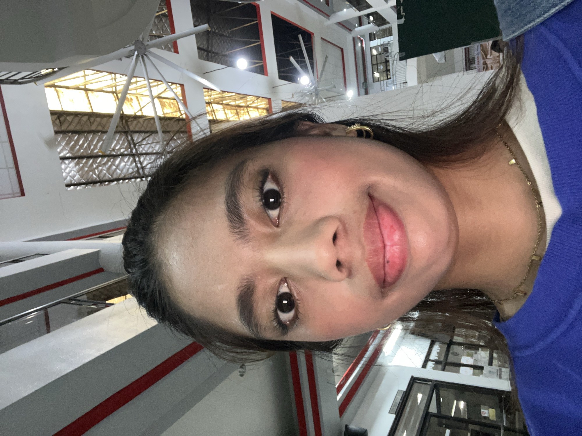 An indoor selfie shows a young woman with dark hair and brown eyes in a close-up shot. She is wearing a blue sweatshirt over a white top and a delicate gold necklace. A small gold hoop earring is visible on her right ear. Her makeup includes blush and lip gloss. The background features architectural elements like diagonal beams with red accents and a ceiling with exposed structural components and lighting fixtures. The overall environment suggests an indoor public space, possibly a mall or a large building, with a bright, airy feel. The provided contextual information states the location is the city of Panabo, Philippines.