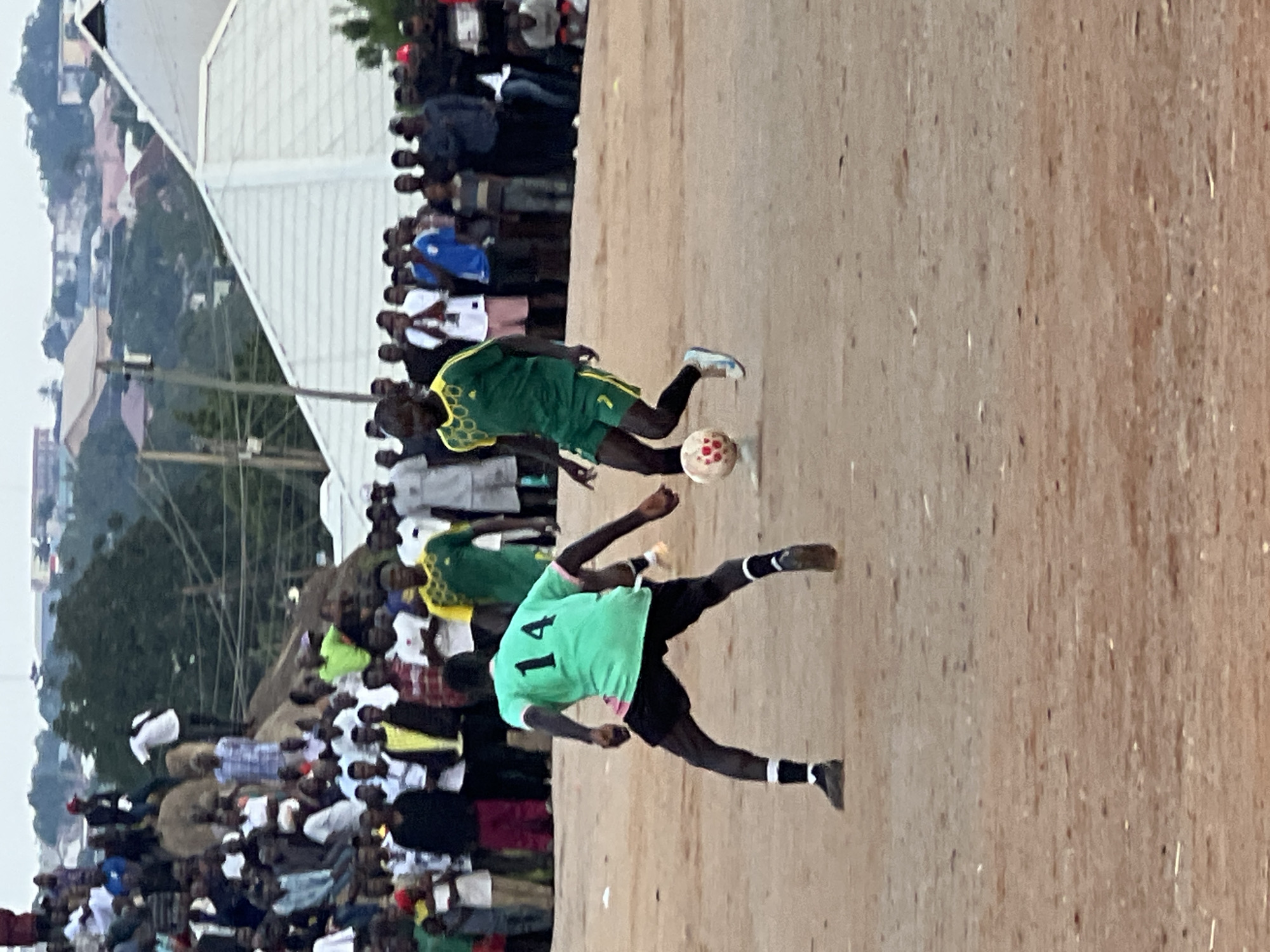 This is a full shot photograph of a soccer game taking place on a dirt field in Jos, Nigeria. The game is being played in a crowded area, with a large group of spectators watching from the sidelines. The spectators are a mix of men, women, and children, and they are all dressed in casual clothing. The field is barren and dusty, with no grass. The players are wearing soccer uniforms, and they are actively engaged in the game. One player in a green jersey with the number 14 on his back is in the foreground, while another player in a green and yellow jersey is to his right. The background of the image shows a dense urban area with buildings and trees. The sky is overcast, and the lighting is soft.