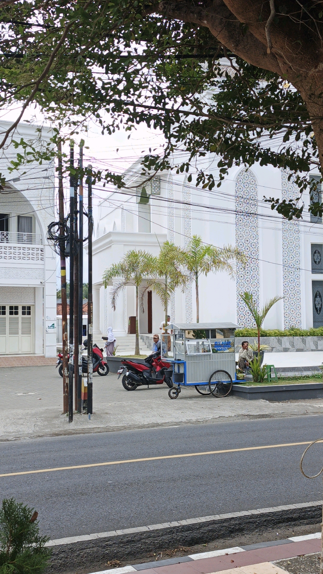 This image captures a street scene in Desa Karangbenda, Indonesia, with a prominent white building, possibly a mosque, in the background. The building features Islamic architectural elements, including arched windows and decorative patterns on its facade. In the foreground, a paved road runs parallel to a sidewalk. Several individuals are present. On the sidewalk, a person is seated on a bench near a cart. Another person is standing in front of the cart. A red motorcycle is parked to the left of the cart, and another red motorcycle is further to the left, partially obscured by utility poles. Several utility poles with numerous wires are positioned in the middle ground. Palm trees are visible in front of the white building, adding to the tropical atmosphere. The sky is overcast, suggesting a cloudy day. The scene appears to be a typical day in a residential or commercial area of Desa Karangbenda.