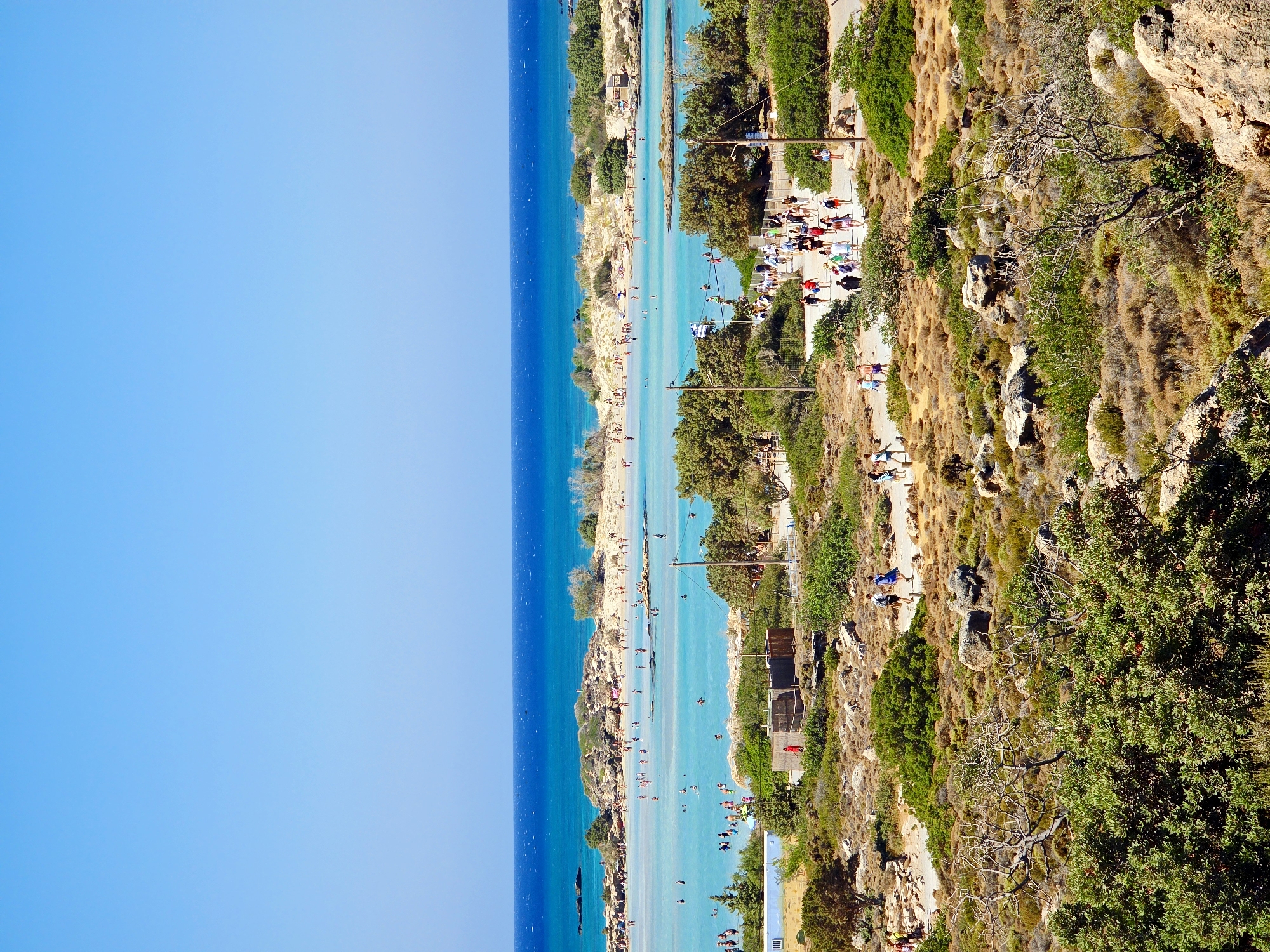 The image shows a wide, scenic view of a tropical beach and turquoise lagoon. In the foreground, there is dry, rocky terrain with scattered green bushes and trees. A group of people are walking on a dirt path. The middle ground features the vibrant blue and green waters of the lagoon, with several small islands and a sandy beach dotted with sunbathers. In the background, the deep blue sea stretches to the horizon under a clear sky. The scene is bustling with activity, with people swimming, walking on the beach, and enjoying the sunny weather.