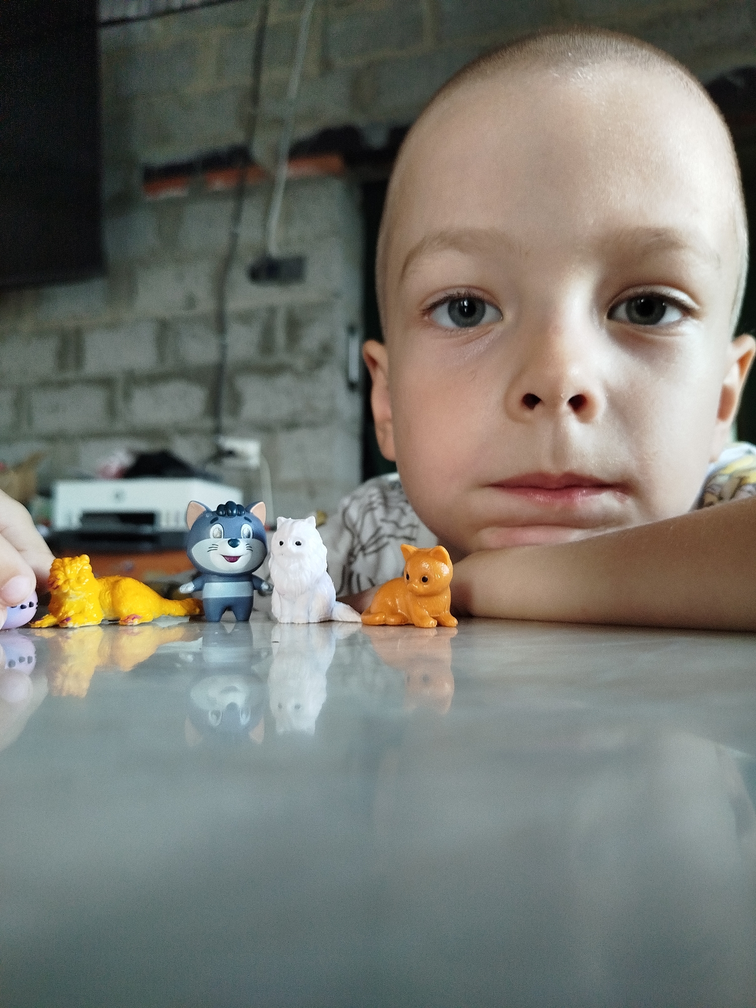 A young boy with a shaved head is looking directly at the camera, resting his chin on his arm. In front of him, on a reflective surface, are several small cat figurines. To the left, there is a yellow cat figurine. Next to it is a grey and white cat figurine with blue stripes. To the right of that is a small white cat figurine. Further to the right is a small orange cat figurine. The background is blurred, showing a brick wall and some electrical wires. The scene appears to be indoors, possibly in a home in Zavodoukovsk, Russia.