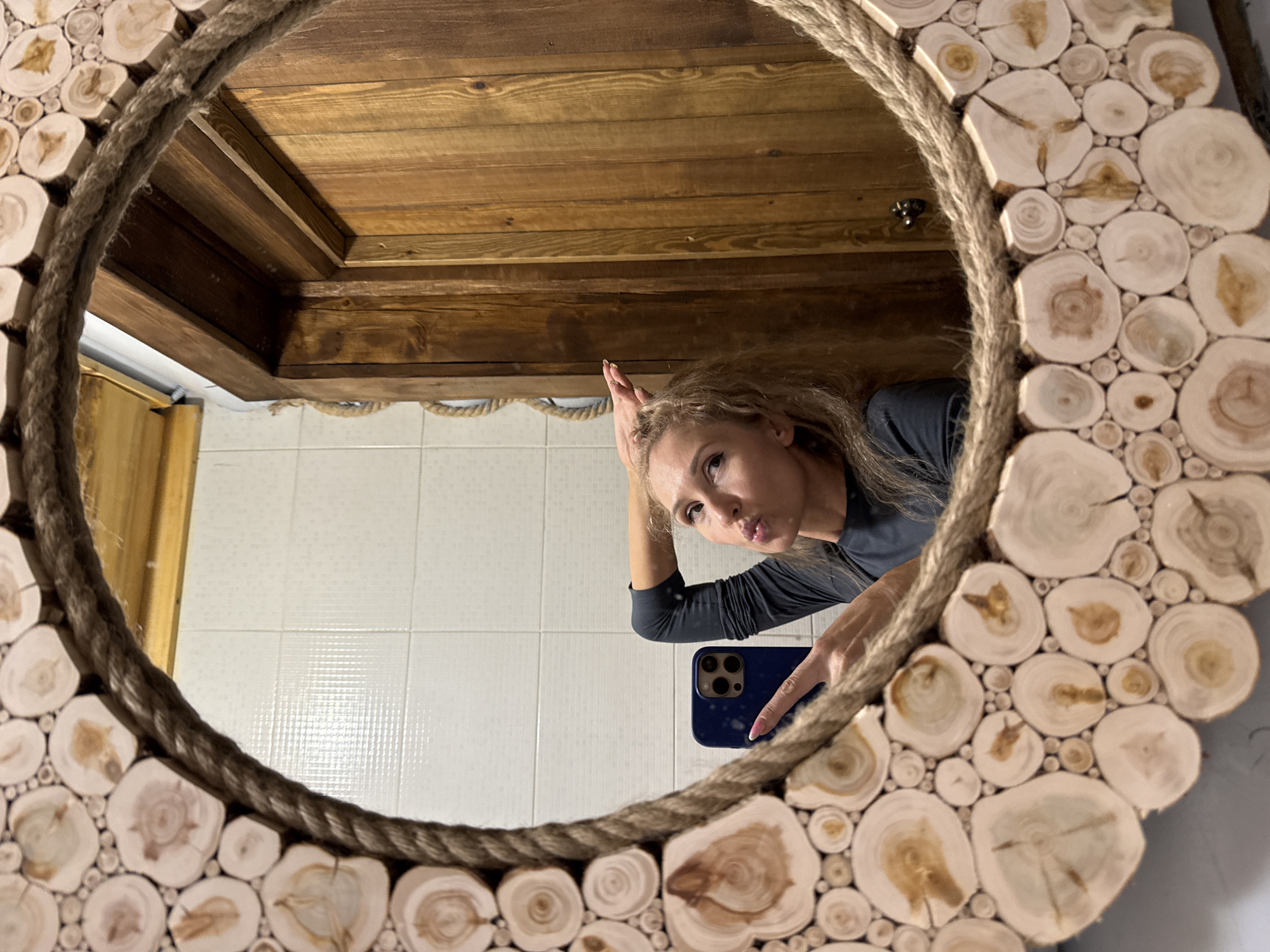 A woman is reflected in a round mirror decorated with rope and wood slices. The mirror hangs on a white tiled wall in a room with a wooden ceiling. The woman is holding a blue cell phone and touching her hair with the other hand. The location is in Perm, Russia.