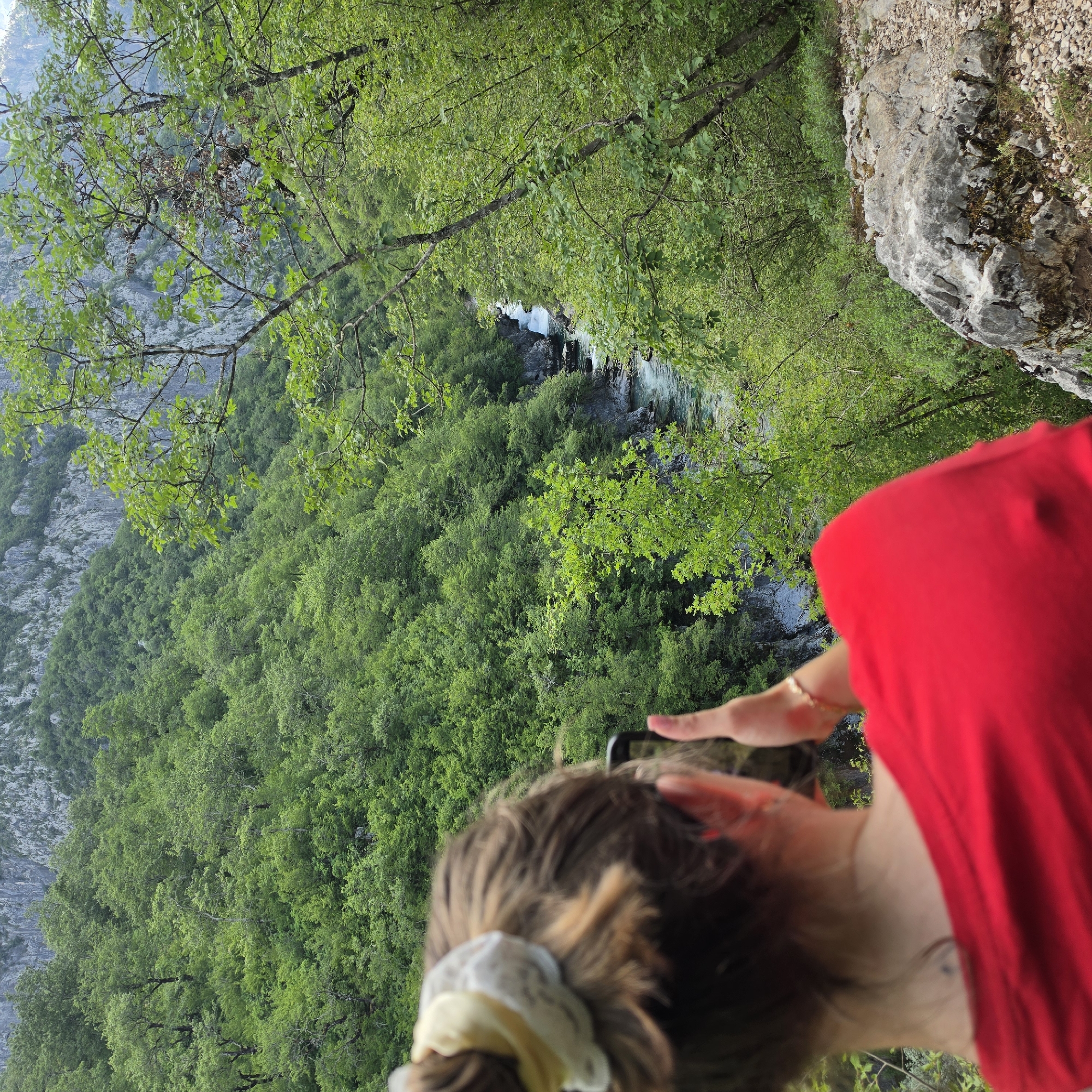 A slightly blurred, outdoor shot shows a person taking a photograph with a cell phone, possibly in Međuriječje, Montenegro. The focus is on the dense green forest and a river flowing through it. To the left, there's a visible mountain face. The person taking the picture is wearing a red shirt and has their hair tied back with a white scrunchie. A rock or cliff edge is visible to the upper right. The person is holding the cell phone in their hands, presumably to capture the landscape.