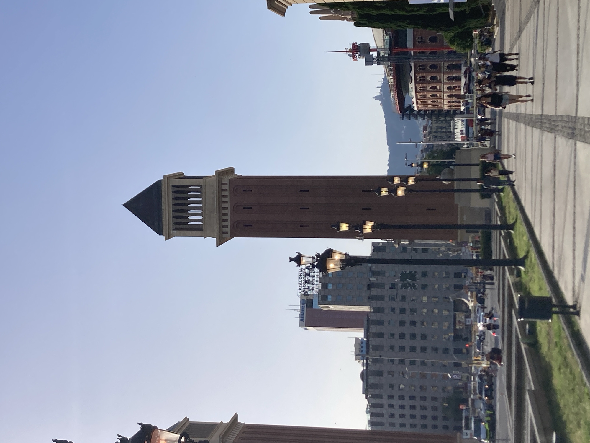 The image depicts a street scene in Barcelona, Spain, likely near the Plaça d'Espanya, with the Venetian Towers (Torres Venecianes) prominently featured in the foreground. The perspective is from ground level, looking down a wide, tree-lined avenue with pedestrian walkways and lampposts.
In the mid-ground, the tall brick Venetian Towers stand on either side of the avenue, flanking a view of distant buildings and a hillside with structures on it. One of the towers has a pyramidal dark roof with decorative openings. The avenue itself is paved with alternating light and dark stones, and several lampposts with multiple lights line its edges.
To the left, a large, grey, multi-story building with a grid-like facade dominates the background, with cars and buses visible on the street in front of it. Further to the right, the distinctive red brick bullring, Arena Barcelona, is visible, with trees and other buildings surrounding it.
Several people are walking along the pedestrian path in the mid-ground and foreground. They are dressed in casual summer clothing, suggesting a warm day. Some are walking individually, while others are in small groups. The overall atmosphere is one of a bustling urban environment on a clear, bright day. The sunlight casts distinct shadows on the ground, indicating it is either morning or late afternoon.
