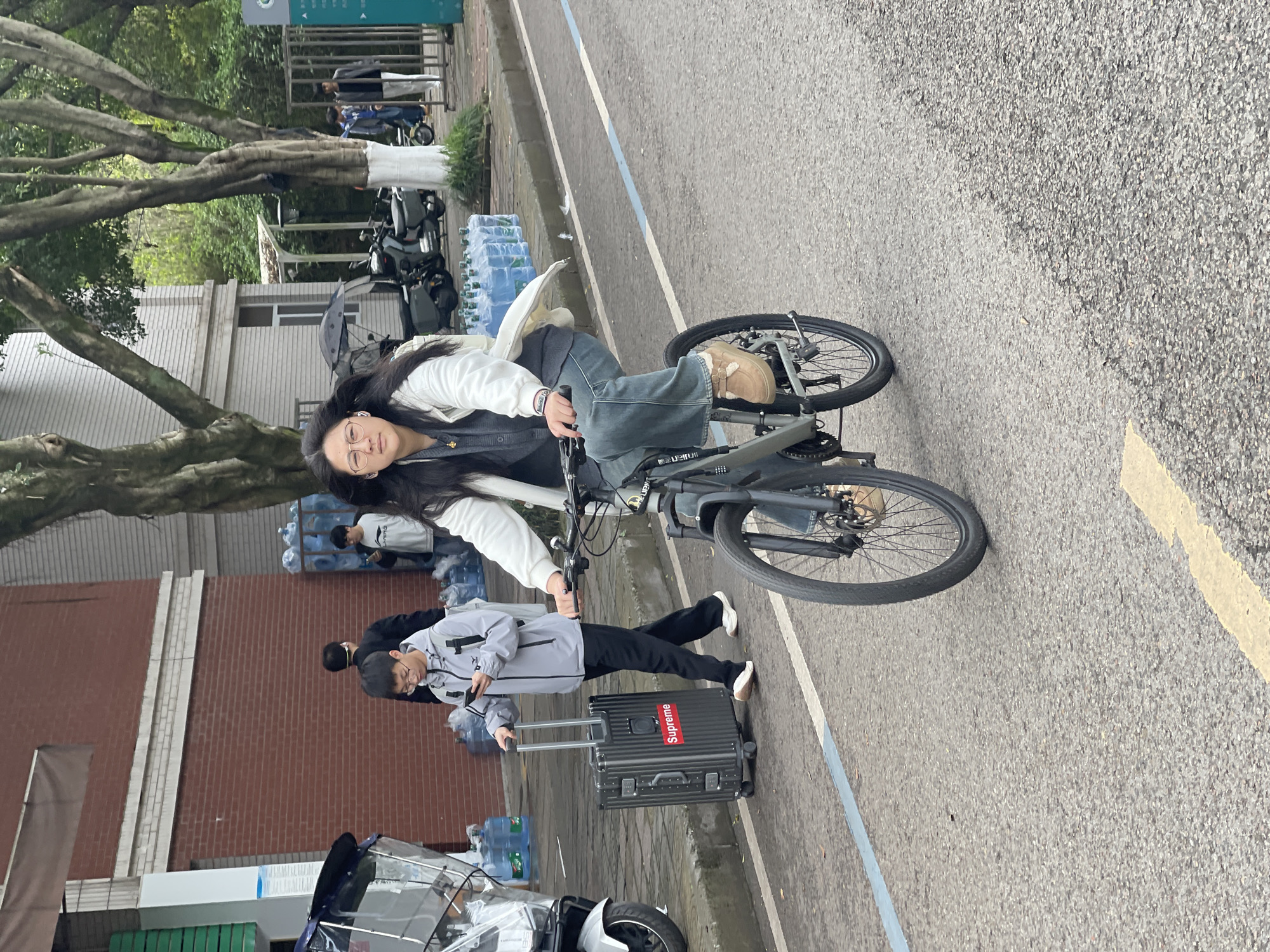 A young woman in glasses, a white sweater, and blue jeans rides a black bicycle on an asphalt road marked with blue and yellow lines. Behind her, a person in a grey jacket pulls a black suitcase with a red "Supreme" logo. To the left, there's a brick building and a large tree. Stacks of water bottles are visible. Other individuals and vehicles are in the background. The location is Chongqing High-tech Zone, China.