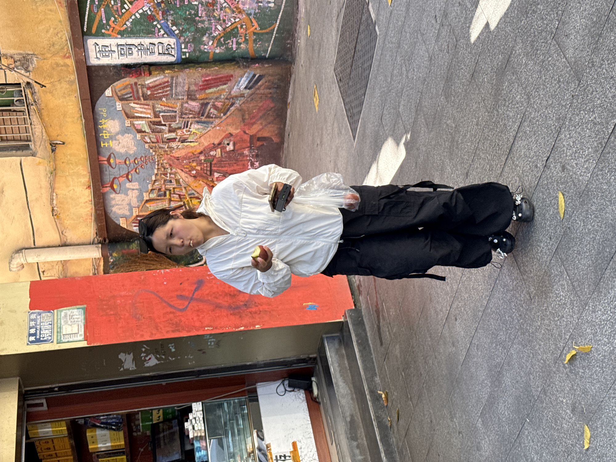 The image shows a woman standing on a gray tiled sidewalk in the Jiulongpo District of China. She is holding a small, green fruit in her left hand, and what appears to be a small electronic device in her right hand. She is wearing a white, lightweight jacket and black, loose-fitting pants with black sneakers. Her hair is dark and pulled back.
Behind the woman is a building with a storefront. The building has an orange-red wall with Chinese characters and other signage. A mural depicting a street lined with books and other objects is visible further up the wall. Part of the store is visible in the lower left corner, including shelves stocked with various products.
The sidewalk also has fallen yellow leaves. The background shows parts of additional buildings with yellow walls.