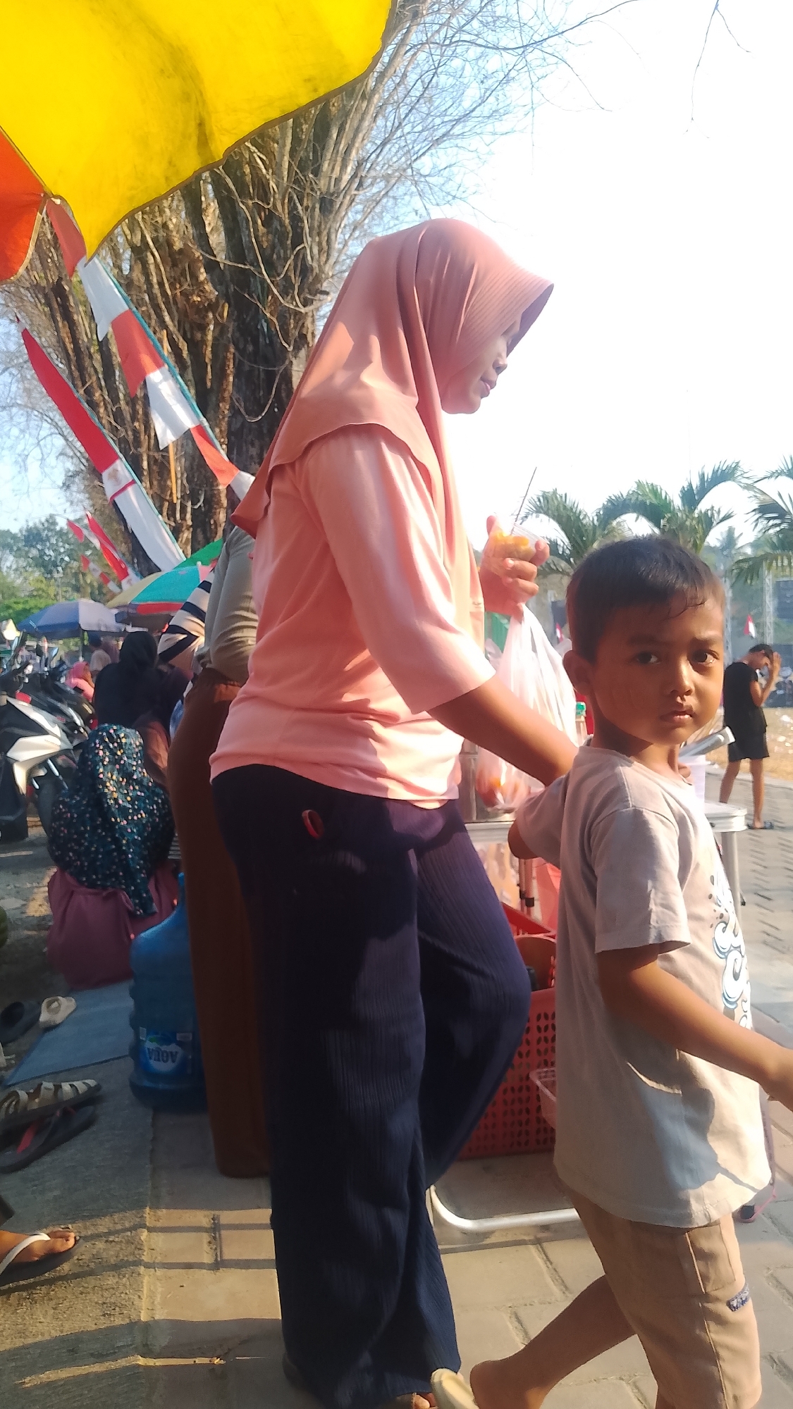 In Karanglangu, Indonesia, a young boy with dark hair and brown eyes stands on a paved walkway, looking towards the camera. He wears a light grey t-shirt with a graphic design and light brown shorts. Beside him, a woman in a peach-colored hijab and matching long-sleeved shirt, along with dark blue pants, walks away from the camera, holding a plastic bag containing what appears to be fruit. In the background, a large tree with bare branches stands tall, partially obscured by a vibrant yellow and red awning. Several parked motorcycles and people in modest clothing are visible, suggesting a market or gathering. A large blue water jug is present on the ground. Palm trees can be seen in the distance. The overall scene is outdoors with bright sunlight casting shadows.