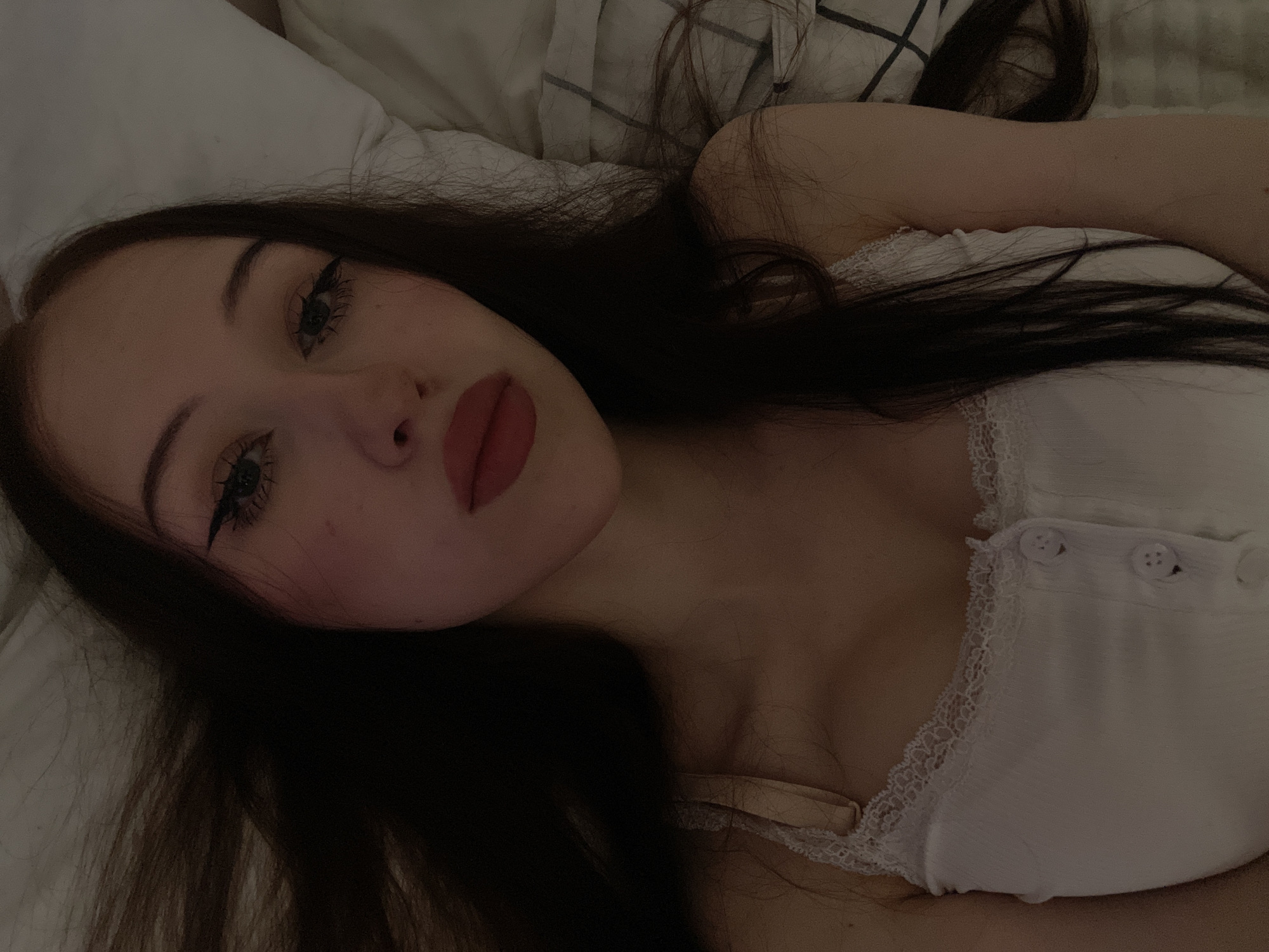 The image shows a close-up, selfie-style shot of a young woman lying in bed, facing the camera. She has long, dark hair that is spread out around her. Her eyes are made up with winged eyeliner, and her lips are painted a deep red. She is wearing a white tank top with lace trim and buttons. The background appears to be a pillow and bedding. The overall lighting is dim, suggesting it might be nighttime or a dimly lit room. There is no indication of the city of Votkinsk, Russia, in the image.