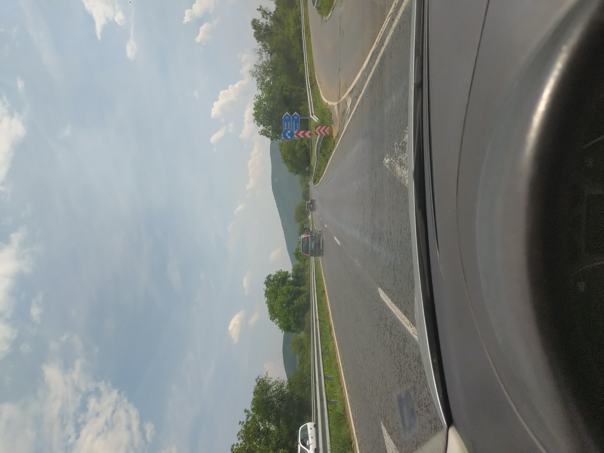 The image depicts a daytime scene from the interior of a vehicle, looking forward through the windshield onto an asphalt road in Zmeyovo, Bulgaria. The road is a two-lane highway with white lane markings, and the surface appears slightly reflective in areas.
In the immediate foreground, the top portion of a dark-colored steering wheel and dashboard of the recording vehicle are visible.
Ahead on the road, a dark-colored SUV with a roof rack is driving away from the viewer. Further in the distance, another dark vehicle is visible. To the left of the main road, on an adjacent road or shoulder, a white van or minivan is partially visible.
A silver metal guardrail runs along the left side of the road, separating it from dense green foliage. The right side of the road features a turn-off or exit. At this junction, a blue directional road sign is prominently displayed. The sign points left towards "ОВО 2" (possibly indicating a destination like Gabrovo or similar, 2 kilometers away) and right towards "МИЛОВО 6" (Milovo, 6 kilometers away). Beneath the sign, a red and white striped chevron warning marker is present.
The surrounding landscape is characterized by abundant green trees and bushes lining both sides of the road. In the mid-ground and background, rolling green hills or mountains are visible. The sky above is light blue with scattered white cumulus clouds, indicating fair weather.
