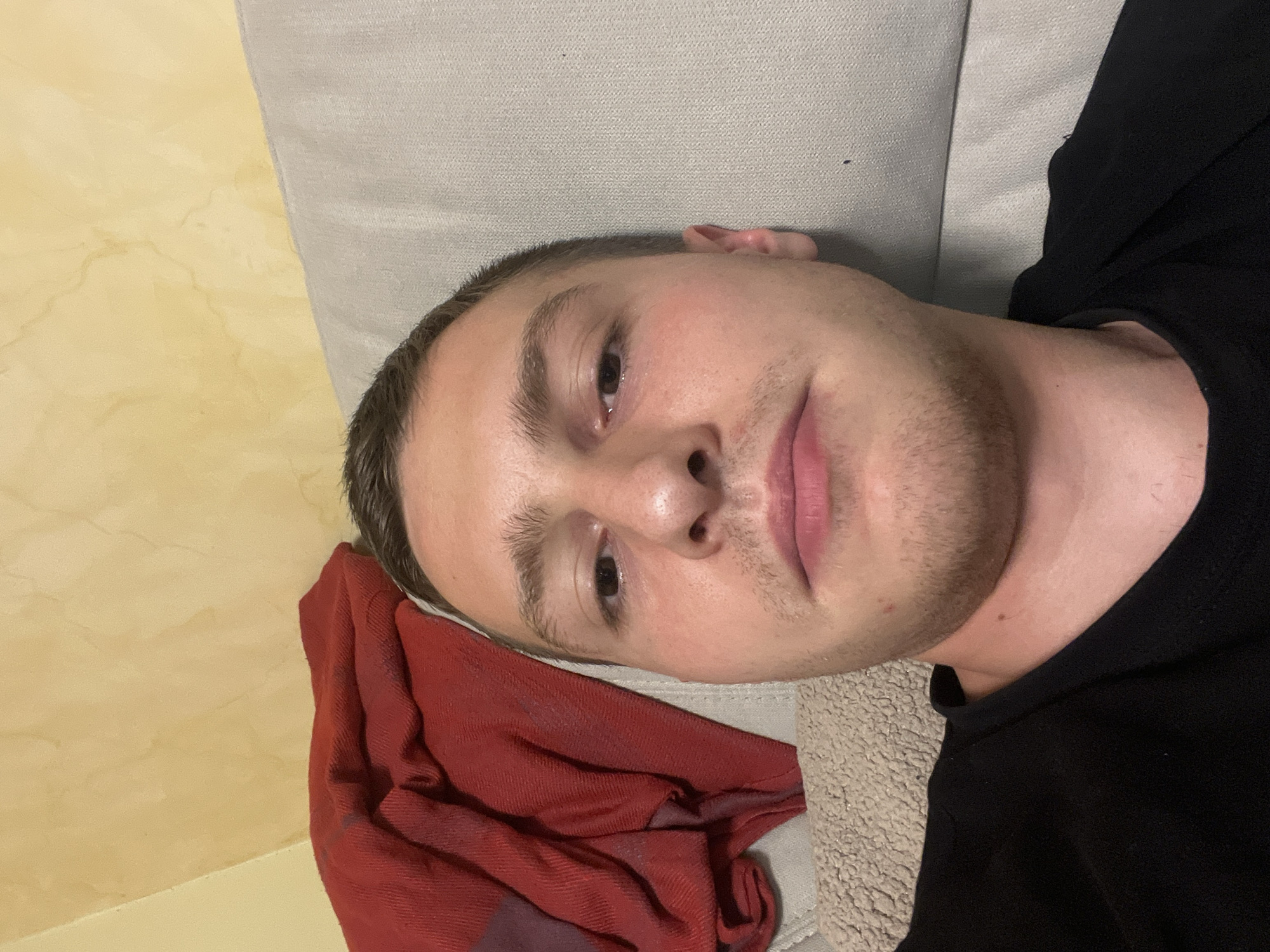 A man with fair skin and short, dark hair is reclining on a light gray couch, wearing a black shirt. His head rests on a crumpled red cloth. The background includes a yellow wall. The location is not discernible in this image and there is no indication it is in Burgas, Bulgaria.