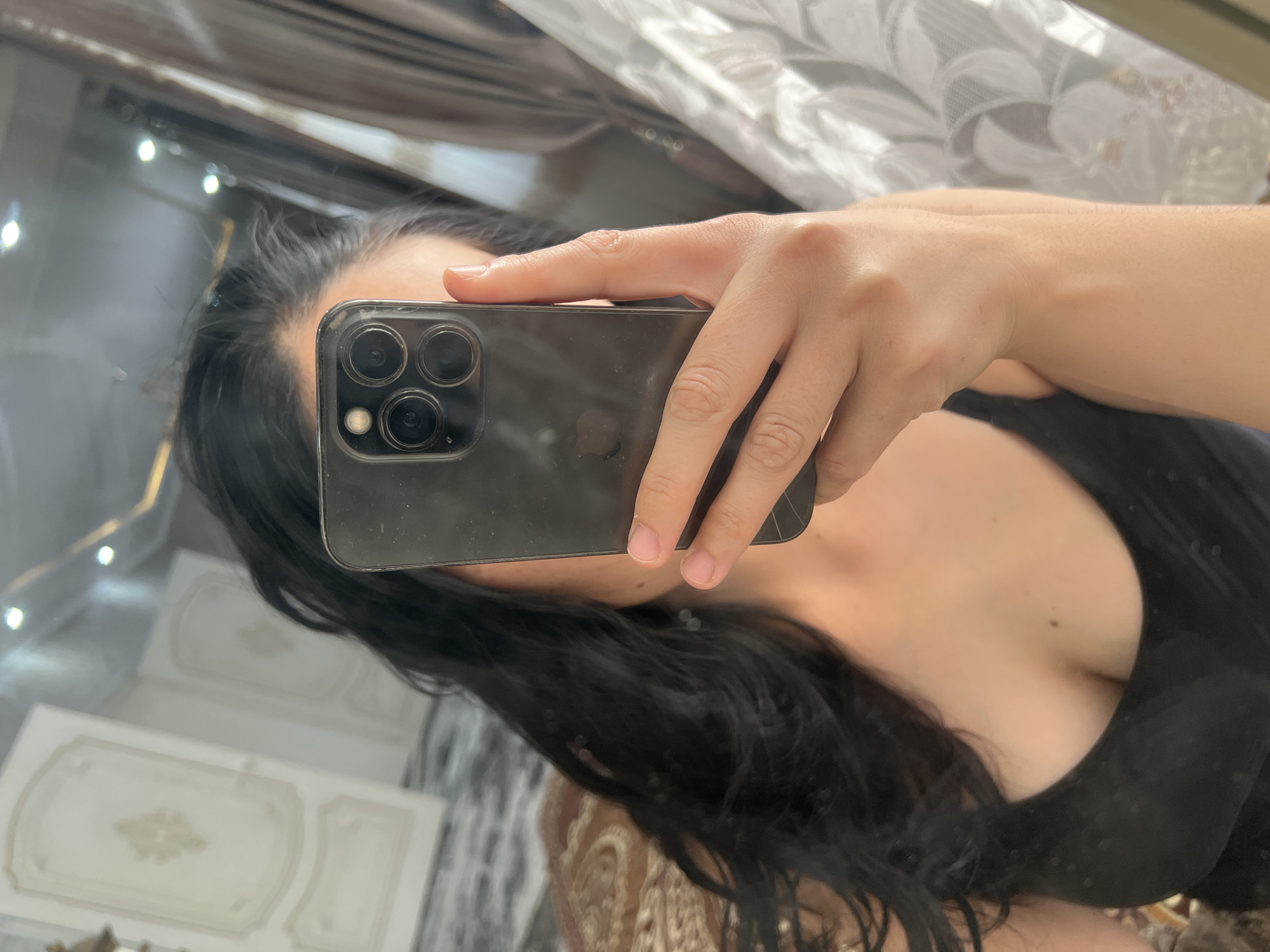 A person is taking a mirror selfie in what appears to be a bedroom in Kara-Dıykan, Kyrgyzstan. The person has long, dark hair and is wearing a black top. They are holding a dark-colored smartphone, which has a prominent camera module with three lenses, in their right hand, angled to capture their reflection. The background is a mirror reflecting a room with ornate wall moldings and a curtain. The lighting suggests it could be daytime, with some artificial lights also visible in the reflection. The overall mood is casual and personal. There is no visible text in the image.