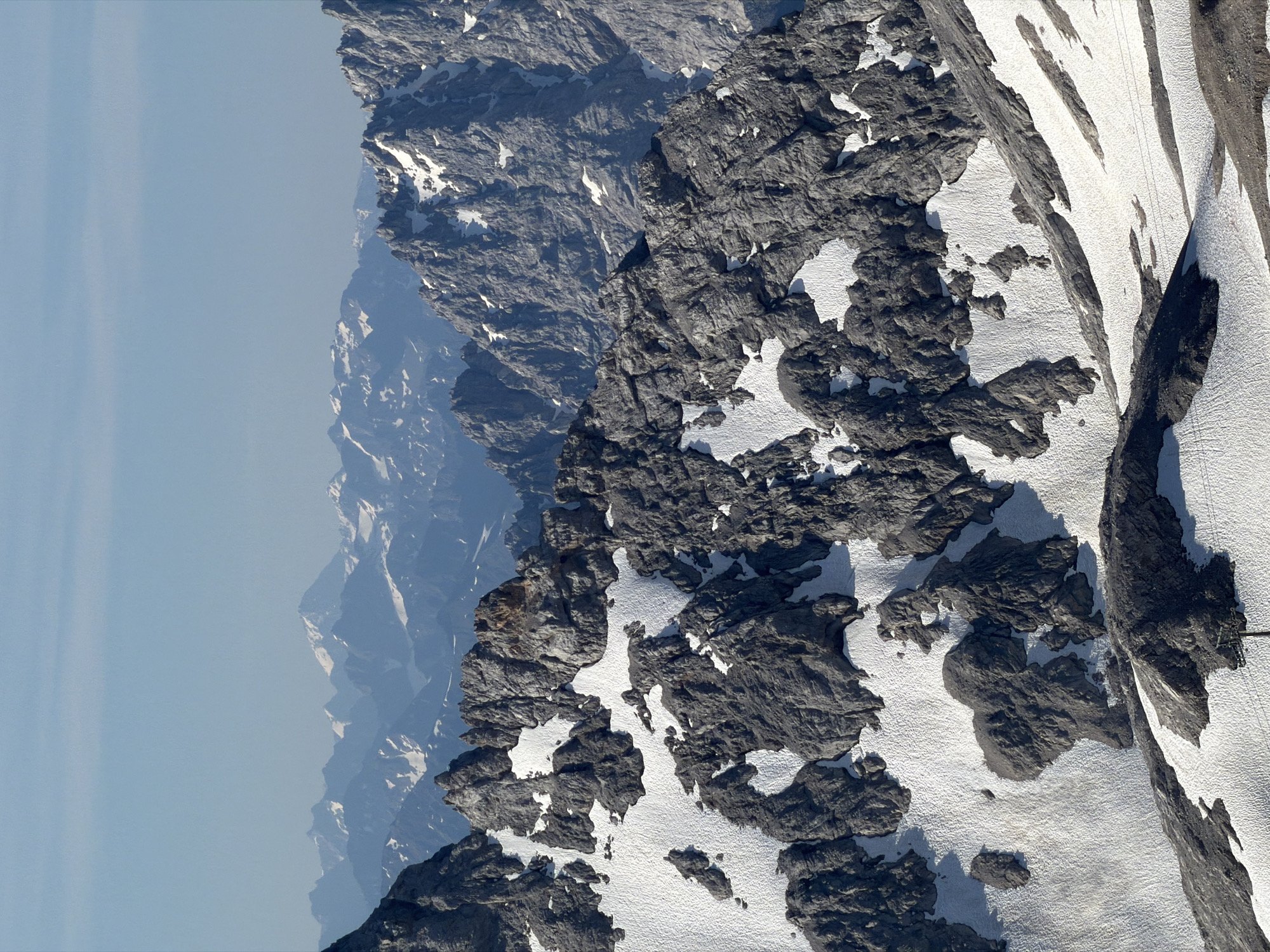 The image depicts a panoramic view of rugged, snow-dusted mountain peaks under a clear blue sky. The foreground is dominated by dark, jagged rock formations interspersed with patches of white snow. In the middle ground, more distant mountains rise, their slopes also partially covered in snow and their rocky textures visible. The background reveals a range of even further mountains, appearing more softened by atmospheric haze and covered in more extensive snowfields. The overall impression is one of a vast, majestic alpine landscape. This scene is characteristic of the Wetterstein Mountains in the vicinity of Garmisch-Partenkirchen, Germany, where the Zugspitze, Germany's highest peak, is located. There are no people or specific man-made objects clearly visible in the main expanse of the image, though a hint of what might be a cable or wire is faintly discernible in the lower right portion.