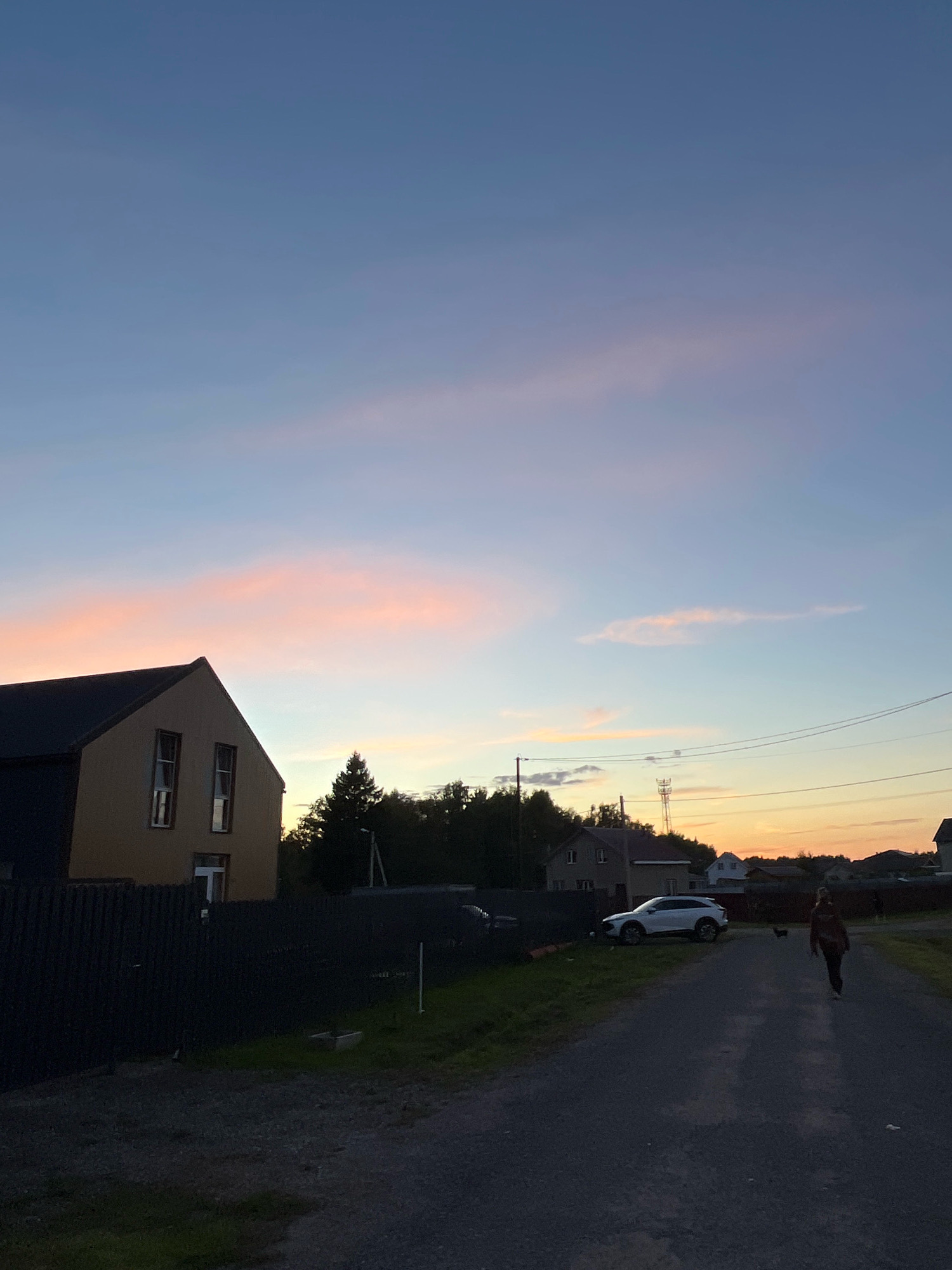 The image depicts a twilight scene in the village of Krasnaya Presnya SNT, Russia. In the foreground, a dark gravel road stretches into the distance. A person is walking away from the camera on this road, accompanied by a small dark animal, possibly a dog. To the left of the road, a dark fence encloses a grassy area. Behind the fence, a modern two-story house with a beige facade and a dark roof is visible, featuring two windows on the upper floor and one on the ground floor. Further back, a line of trees and other houses can be seen. A white SUV is parked on the side of the road near the trees. Power lines crisscross the sky, and a cellular tower is visible in the distance. The sky displays a gradient of colors, transitioning from a pale blue at the top to soft pink and orange hues near the horizon, indicative of sunset.
