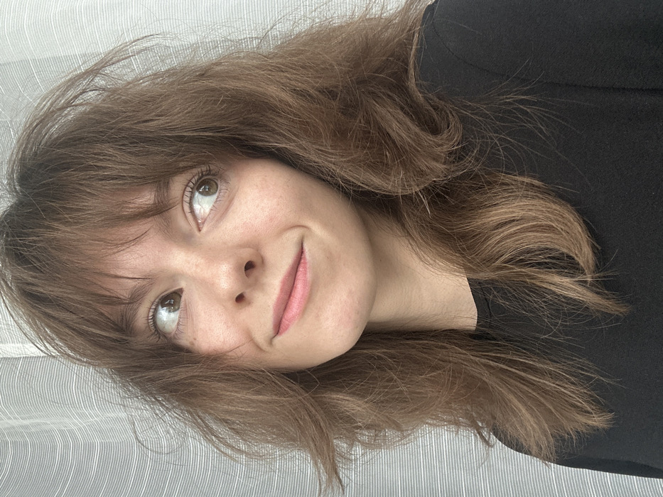 The image depicts a close-up portrait of a young adult female, positioned indoors within the city of Moscow, Russia. She has light skin, brown eyes, and shoulder-length brown hair with lighter highlights and bangs, which appears slightly tousled. A subtle mole is visible on her left cheek. She is wearing a plain black, long-sleeved top. Her head is tilted slightly to her left, and her gaze is directed upwards and to the viewer's right, with a faint, closed-mouth smile indicating a mild, contented expression. The background consists of a light-colored, vertically striped or textured curtain or wall.