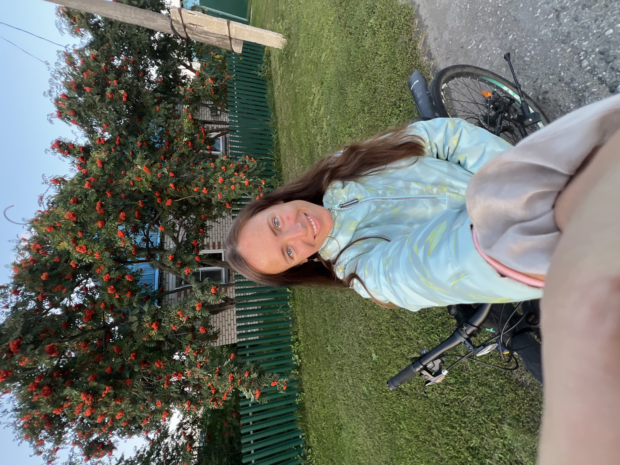In Pivkino, Russia, a woman with long brown hair and blue eyes is taking a selfie. She is wearing a light blue and yellow iridescent jacket and is smiling at the camera. She is standing next to a black bicycle with its handlebars visible in the foreground. Behind her, a rowan tree laden with bright red berries stands tall against a brick house with a green wooden fence. The ground is covered in green grass, and a paved road is visible to the right. The sky is a clear blue, suggesting it is a sunny day.