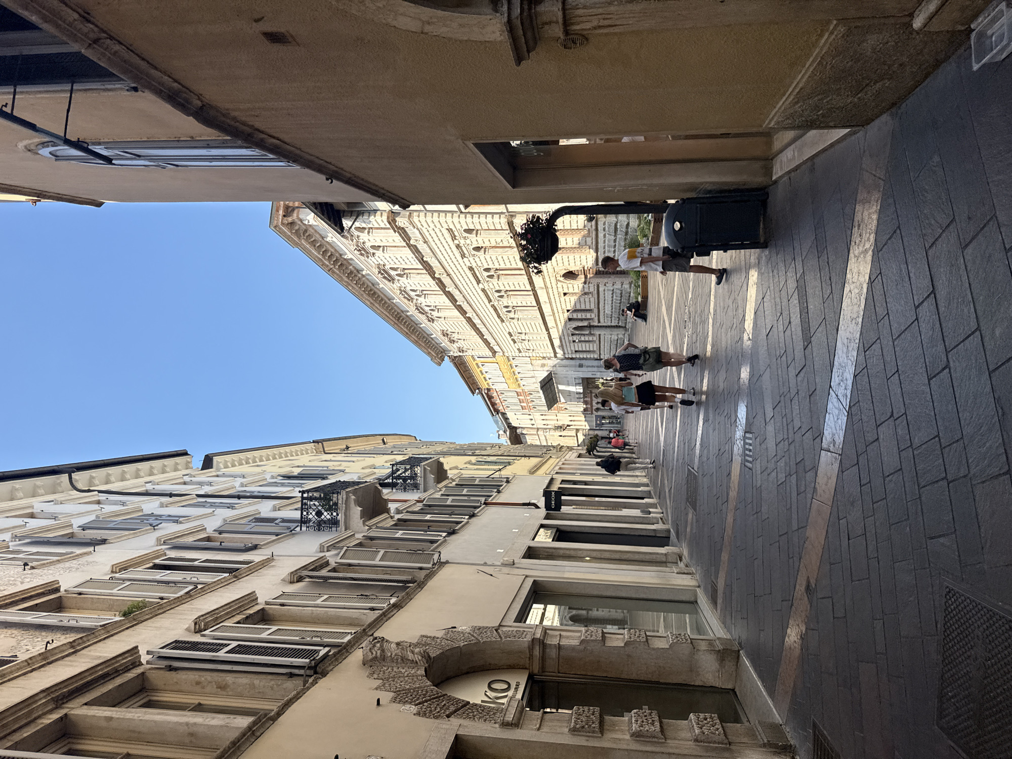 The image shows a narrow street in the historic city center of Trento, Italy, likely on a sunny day with a clear blue sky. The street is paved with dark grey cobblestones, with lighter-colored inlaid stripes forming a pattern. On the left, tall buildings with cream and yellow facades line the street. These buildings feature numerous windows with open shutters, ornate balconies, and architectural details. A shop with a large glass storefront and signage that reads "KO" is visible on the ground floor. Further down the street, more shops with glass windows and awnings are present.
On the right side of the street, a building with a tan facade dominates the foreground. It has a large arched window and a textured lower section. Further down, the street opens up to reveal more buildings with elaborate facades, characterized by rows of windows and decorative elements.
Several people are walking along the street. In the foreground on the right, a young woman with blonde hair wearing a black top and skirt is walking away from the camera. To her left, a young man in a white t-shirt and shorts is walking towards the camera. Further down the street, a group of people are walking in various directions, some appearing to be tourists. One person is seated on a low wall in the distance.
Objects in the scene include a dark green trash bin, a street lamp with a hanging planter, and manhole covers in the pavement. The overall impression is of a vibrant European city street scene on a pleasant day.