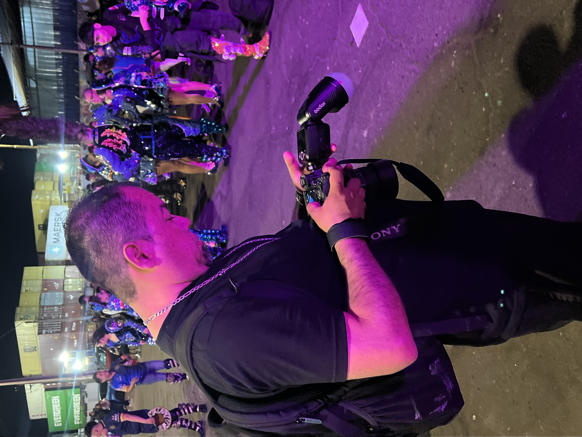 In Arica, Chile, during an outdoor nighttime event, a man in a black t-shirt and backpack is holding a camera with a flash attachment. He is positioned in the foreground, focusing on the camera. In the background, a group of people in colorful, embellished costumes are gathered, some with lights integrated into their attire, suggesting a parade or performance. The environment is illuminated by purple and pink lights, and shipping containers are visible in the background, indicating an industrial or port area of the city.