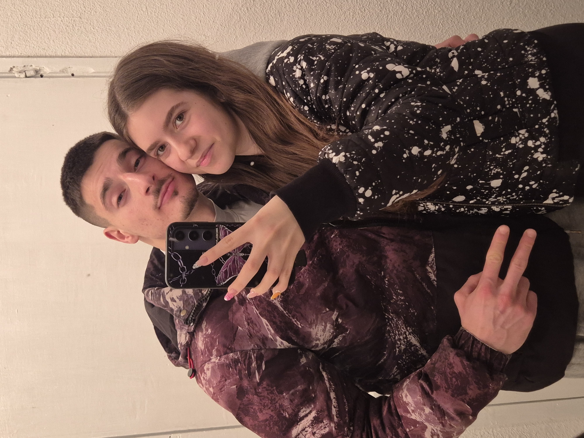 A couple is taking a selfie in what appears to be an indoor setting with a white wall in the background. The man, with short dark hair and a mustache, is holding the phone and appears to be making a peace sign with his left hand. He is wearing a dark jacket with a textured pattern, possibly maroon or purple. The woman has long brown hair and is leaning on the man's head, smiling at the camera. She's wearing a black and white speckled hooded jacket. The camera phone has a dark case with what seems to be a design featuring chains and a butterfly. The woman's nails are painted and have a decorative design. It is impossible to place the location of this photograph in Pavlikeni, Bulgaria based on the given image.
