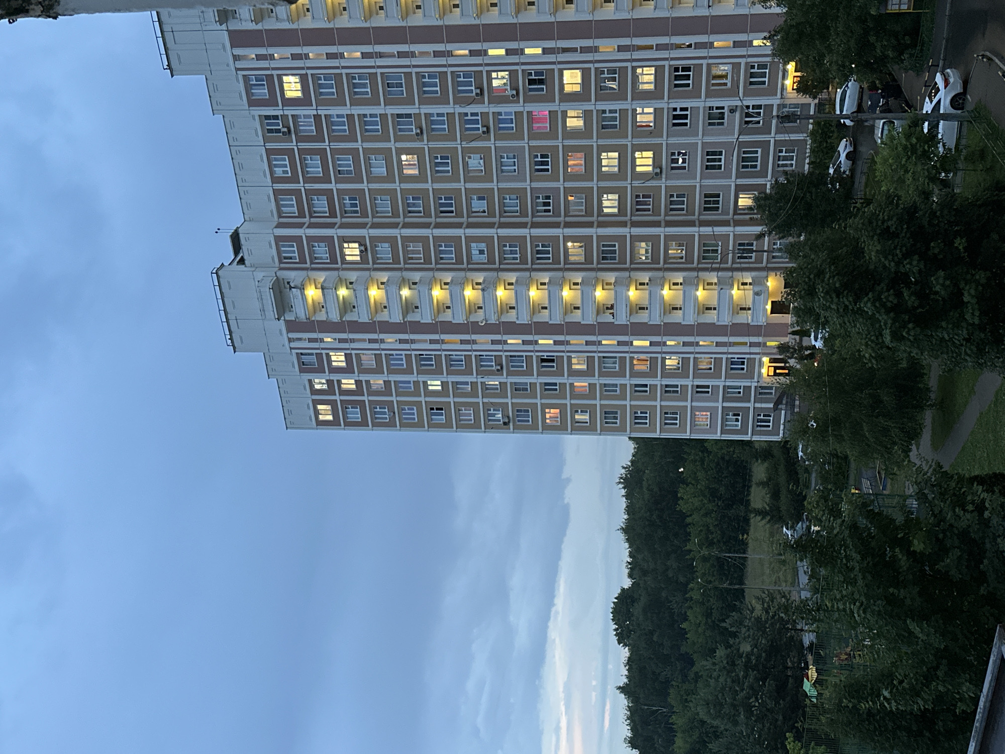 The image depicts a tall, multi-story apartment building in Podolsk, Russia, at dusk. The building, with a cream-colored facade and pink vertical accents, features numerous windows, many of which are illuminated with a warm, yellow light, suggesting residents are home. Balconies with railings are visible on some floors.
In the foreground and to the left of the building, dense green trees and foliage dominate the lower portion of the frame. A playground area with green fencing and some play structures is partially visible amidst the greenery. Adjacent to the building and the trees, a paved area serves as a street or parking lot where several cars are parked, including white sedans and a darker vehicle. Streetlights are visible, casting a faint glow.
The sky above is a muted blue, with visible clouds contributing to the twilight atmosphere. The overall scene suggests a residential urban environment in Russia during the evening hours.