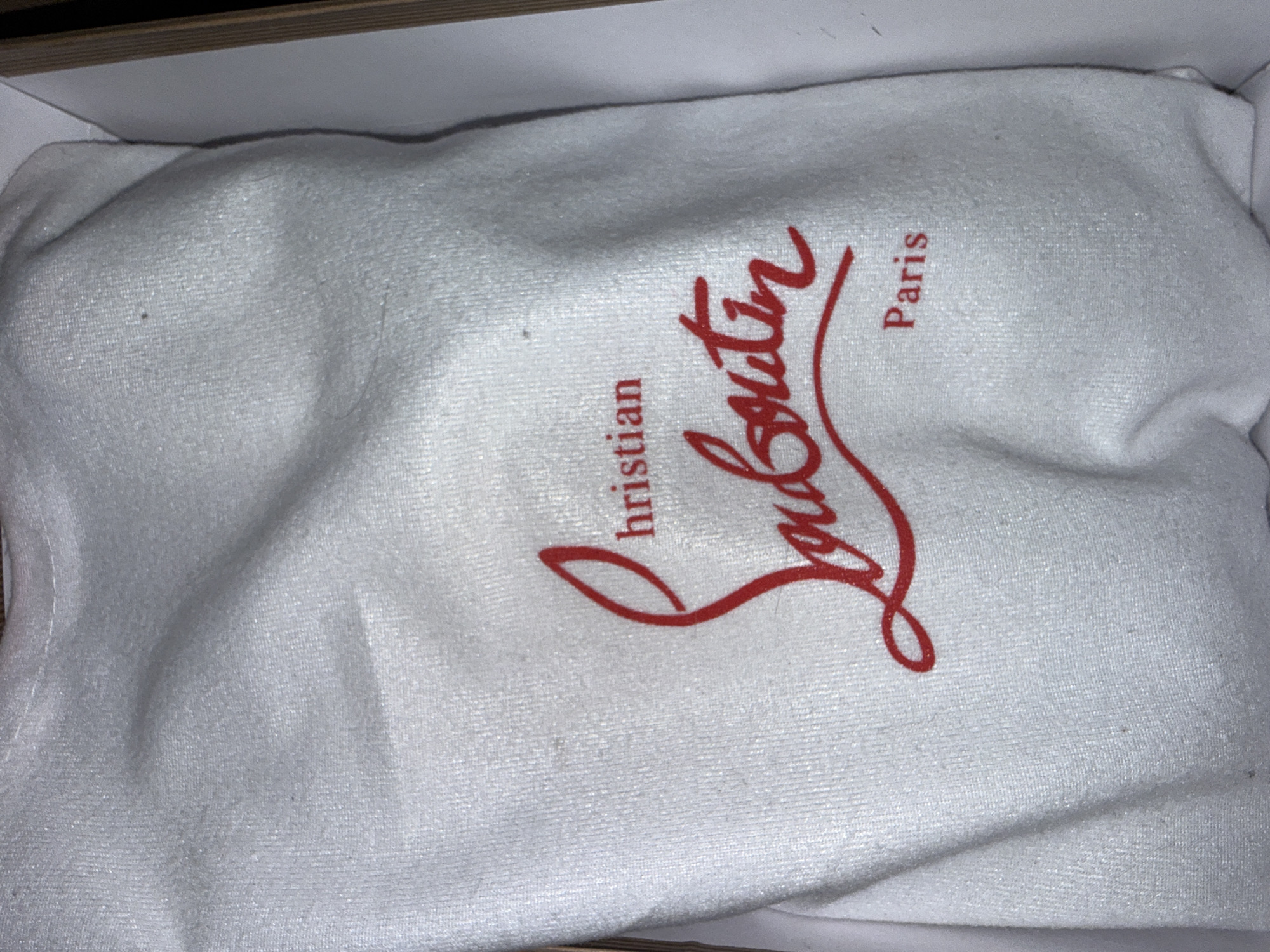 The image shows a white cloth dust bag with a red logo and text. The logo is a stylized script font, with the word "Christian" above a larger script word that appears to be "Louboutin". Below "Louboutin" is the word "Paris". The dust bag is inside a box. The image is a close-up of the dust bag, with no discernible city or environmental context.