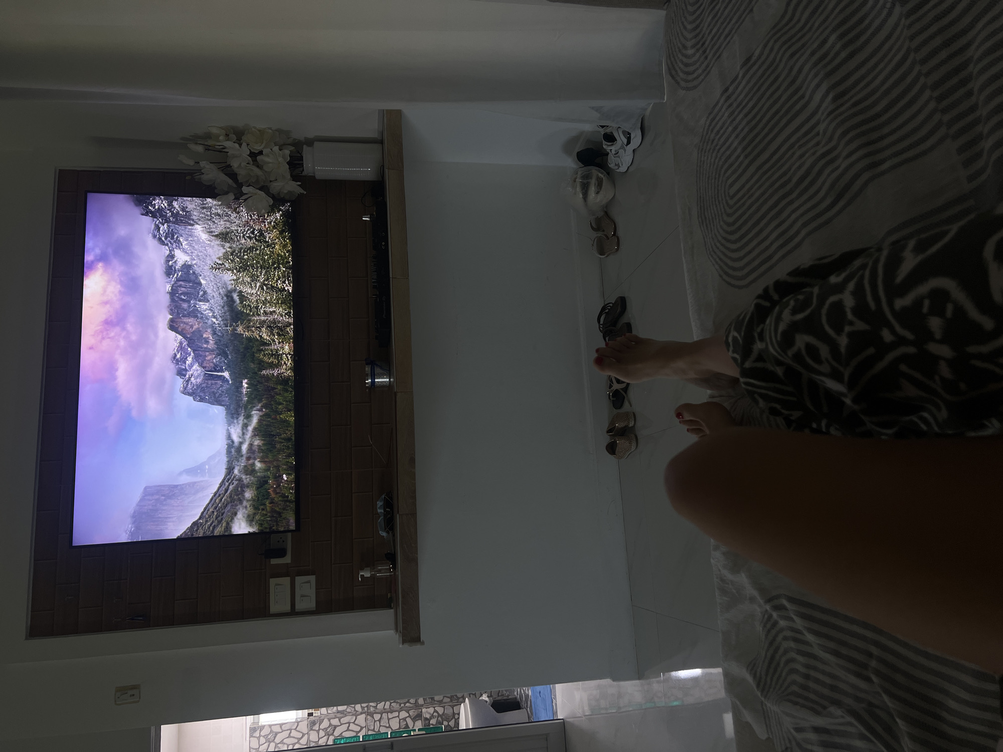A person is lying on a bed in a dimly lit room, looking at a large television screen mounted on a brick-patterned wall. The television displays a scenic image of snowy mountains and a waterfall, with a forest in the foreground. Several pairs of footwear are scattered on the tiled floor near the wall, including sandals and sneakers. In the background, a doorway leads to another room with tiled walls and a toilet visible. The bed is covered with a striped duvet.