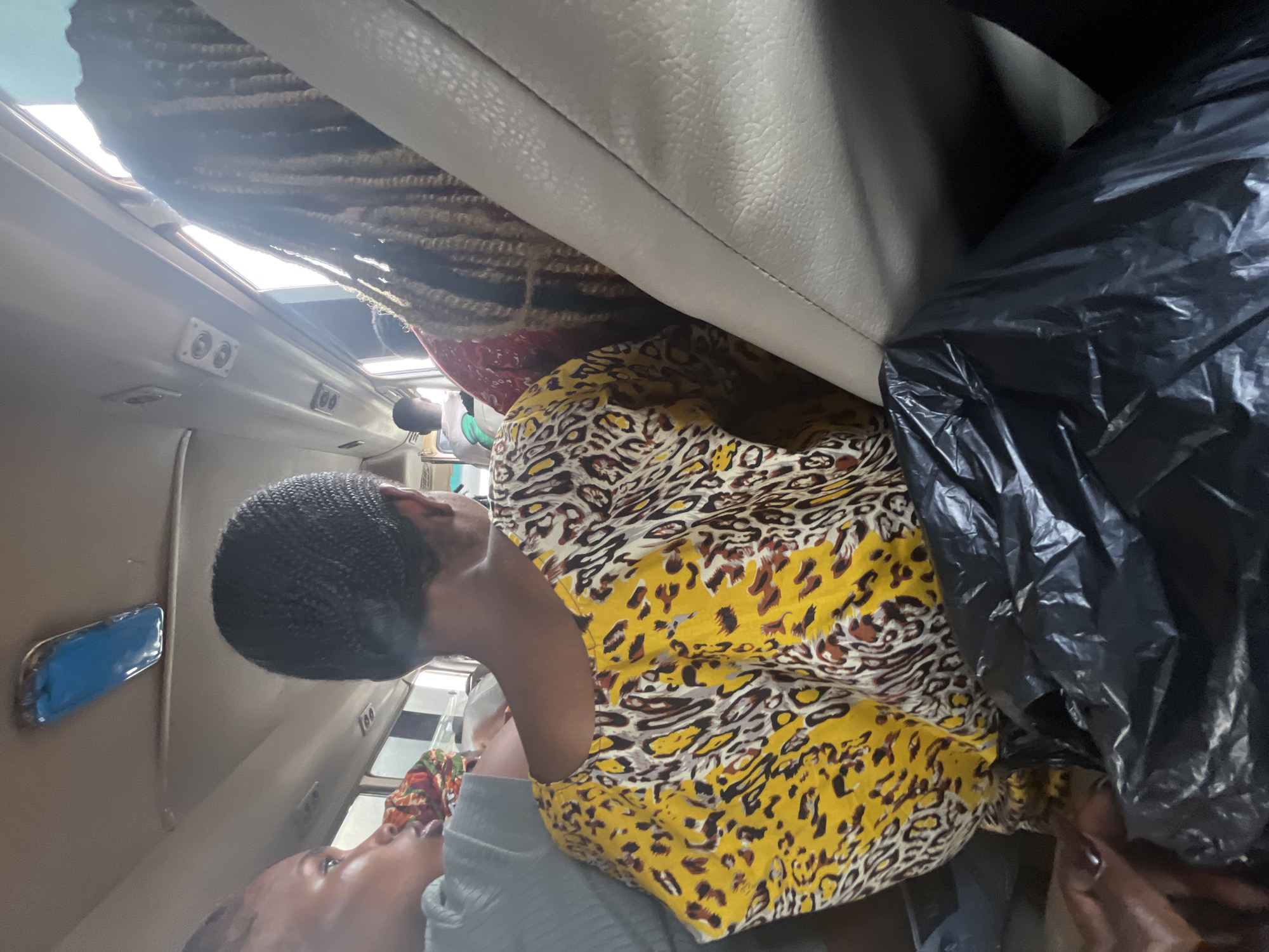 The image shows two women inside what appears to be a bus or van, with luggage and a black trash bag also present. One woman in the foreground is wearing a yellow and black leopard print top and a black braided head covering. She is looking downwards, with her face partially obscured by her hand. The other woman is visible in the lower left corner, looking upwards with her mouth open. She is wearing a gray top. The interior of the vehicle is visible, including seats, a window, and light fixtures on the ceiling. The lighting suggests it might be daytime, possibly overcast, as the interior is somewhat dim. The scene appears to be in motion, given the context of being inside a vehicle. The luggage and trash bag suggest travel or the aftermath of a journey. There are no visible signs of text in the image. The setting is Kugbo, Nigeria.