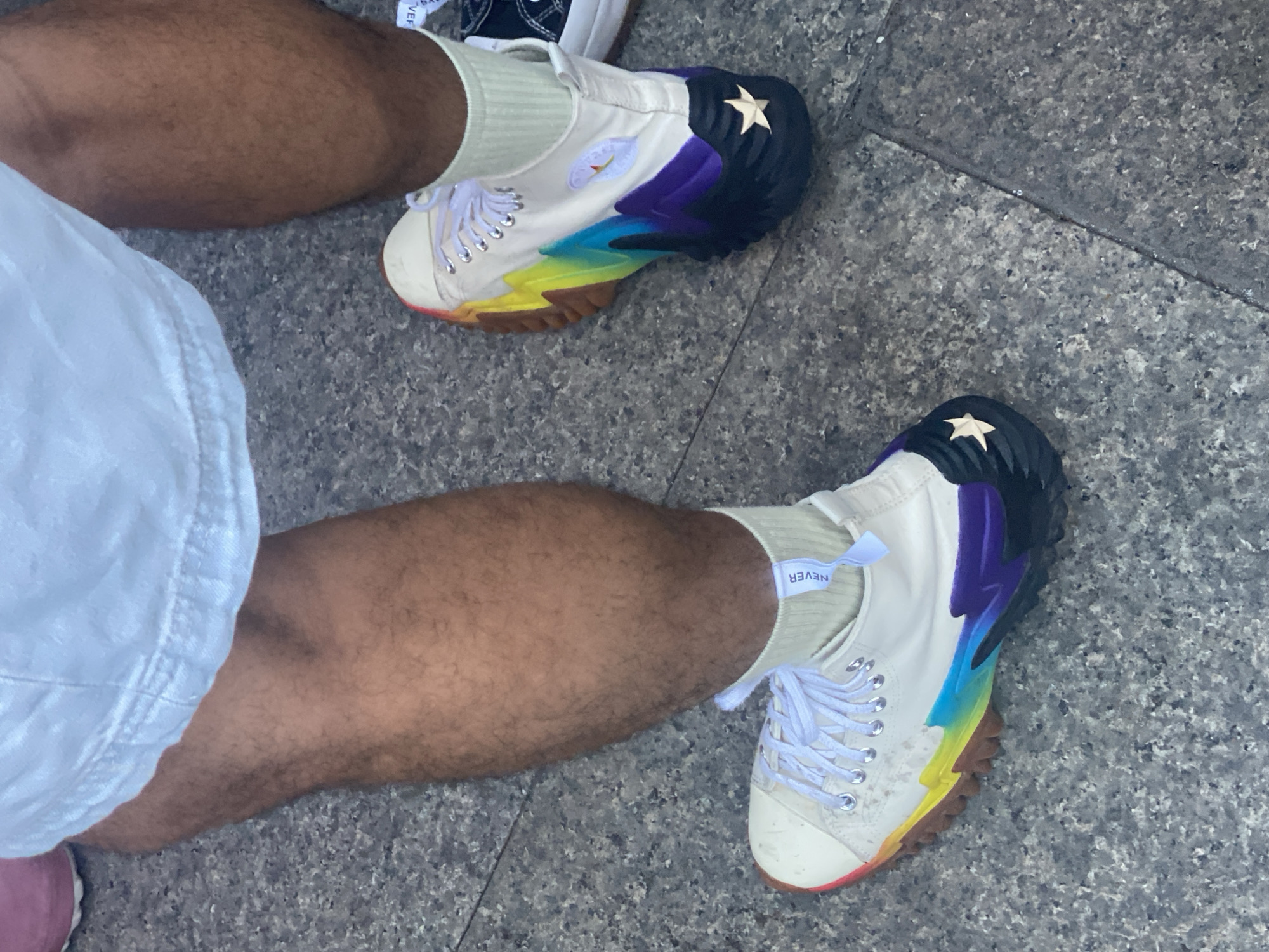 The image shows a close-up view of a person's legs and feet wearing colorful high-top sneakers. The shoes have a white base with rainbow-colored accents on the soles, featuring shades of yellow, orange, red, blue, purple, and black. A small gold star is visible on the heel of the right shoe. The person is wearing light-colored socks and white shorts. The background appears to be a grey, speckled surface, possibly pavement or granite tiles, consistent with an urban outdoor setting. The specific location in Madrid, Spain, is not identifiable from the visual information provided.