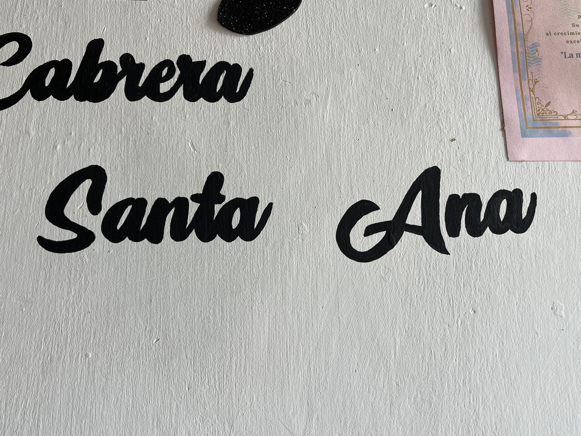 The image displays a white textured wall with black cursive lettering. The words "Cabrera" are written across the top, with "Santa Ana" written below. The lettering is stylized and appears to be hand-painted or applied as decals. The background is a plain white wall with visible texture. To the right of the lettering, a small portion of a pink certificate or diploma with gold and blue decorative borders is visible. The certificate has some Spanish text printed on it. The scene is indoors, and the lighting suggests a well-lit environment. The image does not contain any people or explicit indications of a specific location within Santa Ana, Venezuela, beyond the name itself being written in the image.