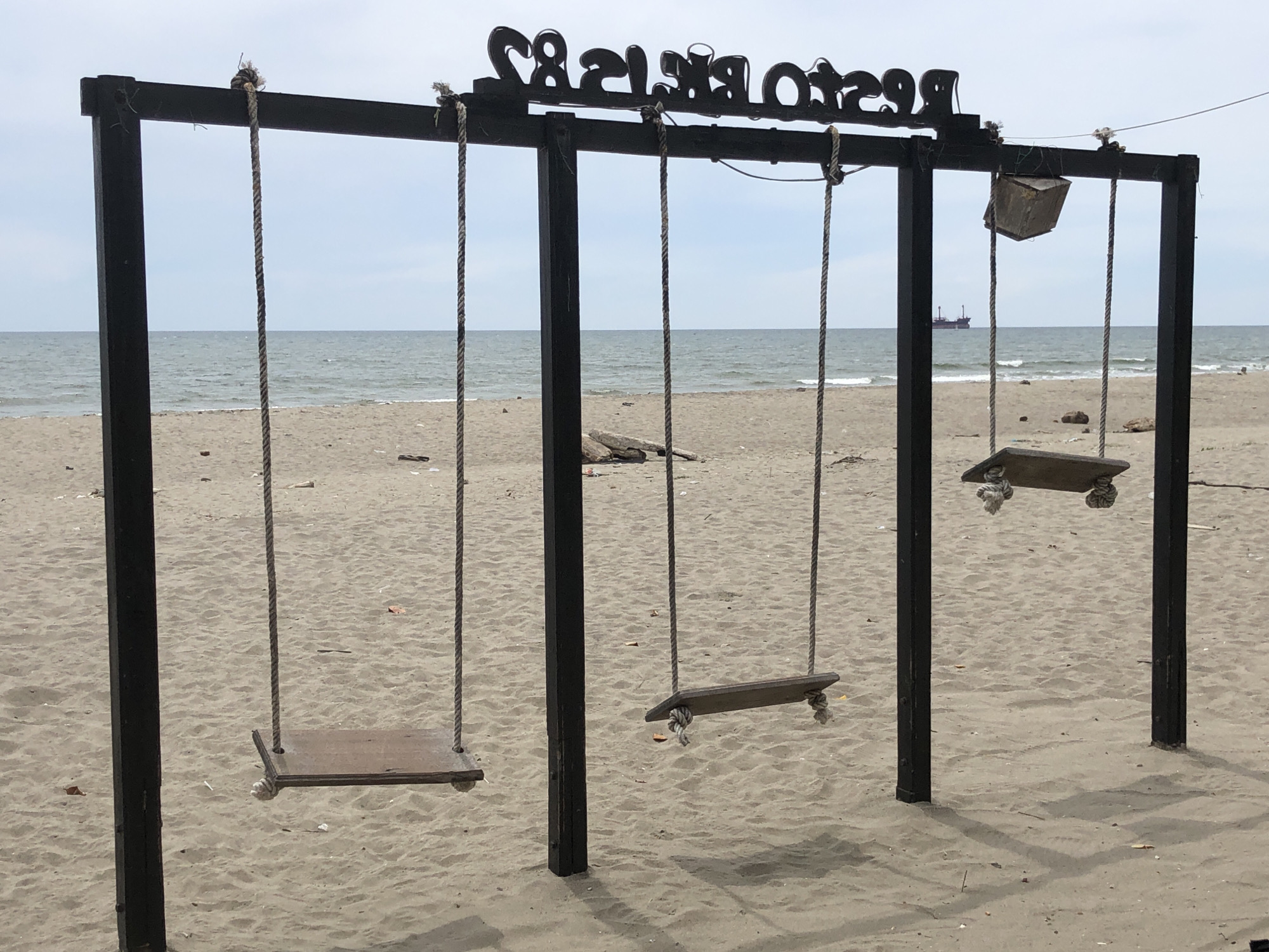 This media file depicts a swingset on a sandy beach with the ocean in the background. The swingset is constructed of black metal posts and a crossbeam. Three wooden swings are suspended by ropes. One swing on the far right appears to be made from a single plank of wood, while the other two are more traditional plank seats. Above the swings, a black sign with white lettering is affixed to the crossbeam. The lettering is in a decorative font and reads "RESTAURANT & BAR." In the distance, a large ship is visible on the water. The sky is overcast, suggesting a cloudy day. The sand is light brown and shows some footprints and debris. The overall scene conveys a relaxed, beachside atmosphere. The location is identified as Keudekruenggeukueh, Indonesia. There are no people visible in the media file. The time of day appears to be daytime due to the natural light illuminating the scene.