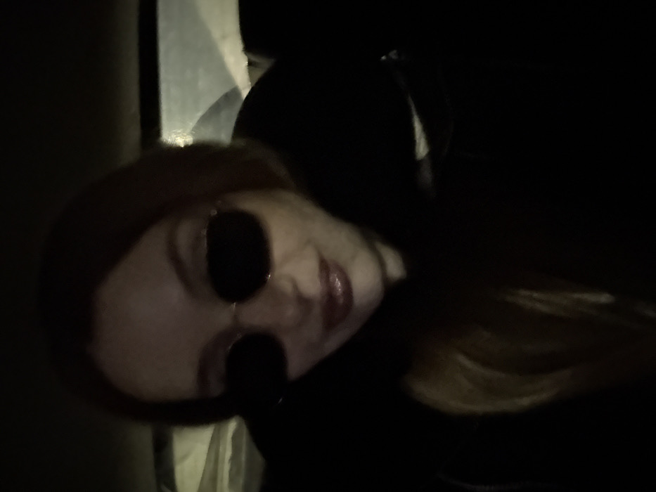 A single individual, an adult female with long, light brown hair, is depicted in a close-up, slightly upward-angled shot. She wears dark, oval-shaped sunglasses with thin metal frames and a dark, possibly black, hooded jacket or coat. Her lips are visible, coated with a glossy, light red or pink lipstick, and she exhibits a slight smile. The scene is dimly lit, suggesting nighttime or a low-light interior environment, possibly inside a vehicle due to the indistinct background features, which include what appears to be a blurred window or light source in the upper right. No visual information within the image definitively identifies the location as Sozina, Montenegro. The individual is looking directly towards the camera, suggesting a selfie or a posed photograph.