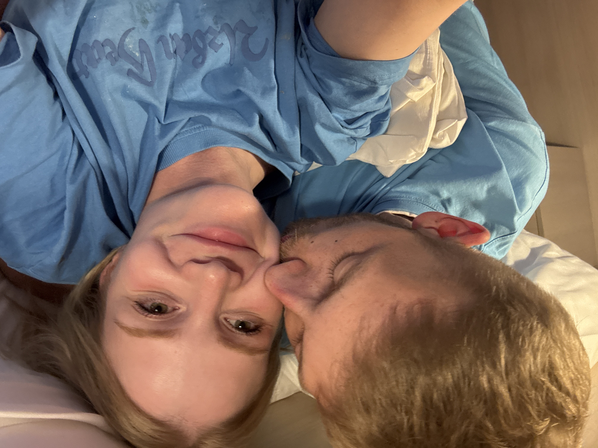 The media file is an upside-down, close-up, eye-level selfie of two people, a woman and a man, lying in bed. The woman is on the left, with fair skin, blonde hair, and green eyes. She is wearing a light blue t-shirt with some dark blue text on it, which is partially obscured. The text appears to be in a stylized font and reads something like "King Size" or "King State". The man is on the right, with fair skin and blonde hair. He is leaning in and kissing the woman on the cheek. He is also wearing a light blue t-shirt, similar to the woman's. The setting appears to be a bedroom, likely in Saint Petersburg, Russia, given the provided context. The lighting is dim, suggesting it is either early morning or nighttime. The overall emotion conveyed is one of affection and intimacy. There are no other discernible objects or environmental details in the frame.