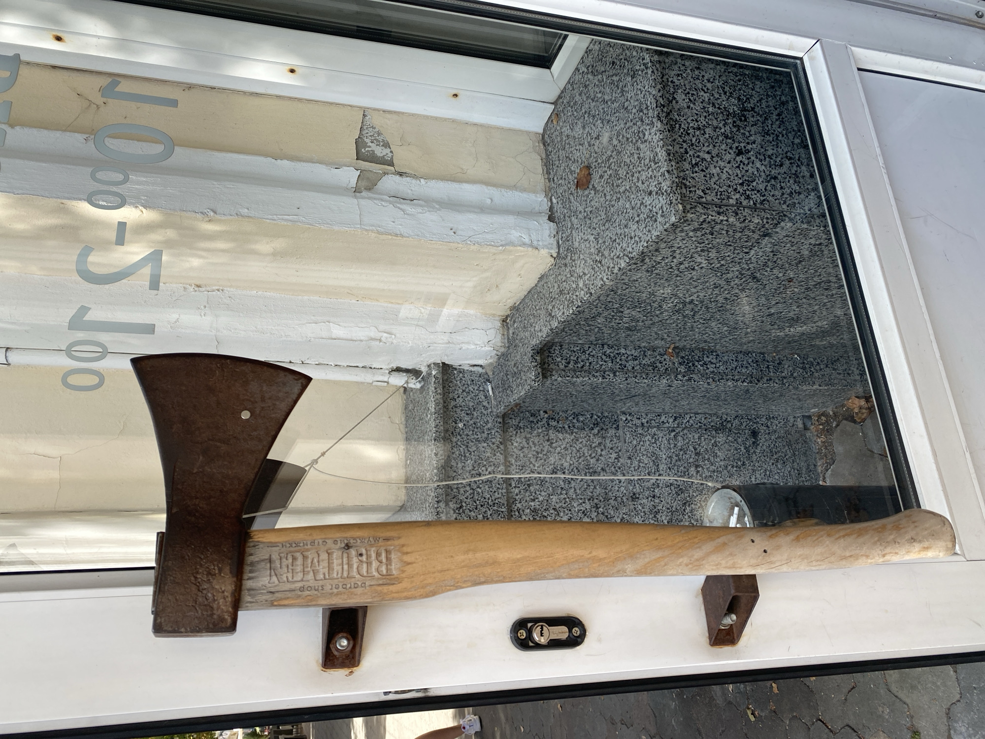 An axe is mounted vertically on the exterior of a white door, which appears to be the entrance to a business. The axe head is rusty and made of metal, while the handle is wooden and shows the branding "BRUMBY" and "barber shop - мужские стрижки" (barber shop - men's haircuts). The axe is secured to the door by a rusty metal bracket near the top and a similar bracket near the bottom. The door has a visible keyhole lock.
The door is positioned in front of a building with a light yellow and white facade. The building has a stone ledge made of granite with a speckled pattern. Reflected in the glass of the door are painted numbers and text that appear to indicate business hours, possibly "09-00" or "09-20". Part of a person's legs, wearing white sneakers and shorts, are visible to the left of the door, suggesting the photograph was taken by a passerby. The environment suggests an urban setting, with a paved walkway visible in the background. This scene is located in Poltava, Ukraine, a detail inferred from the Cyrillic text on the axe handle.
