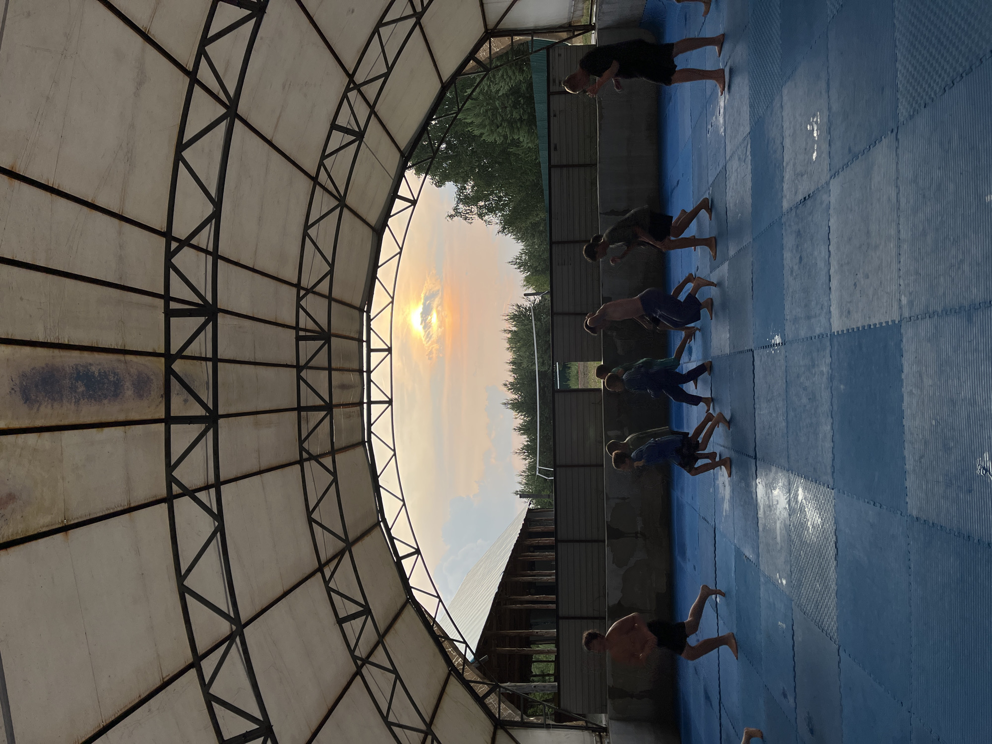 In Krasnoturansk, Russia, a group of young men are engaged in a vigorous outdoor workout session under a large, open-air canopy with a distinctive metal framework. The canopy's translucent white covering allows the warm, diffused light of a sunset to illuminate the scene. The sky is a canvas of soft orange and yellow hues, with the sun peeking through a layer of clouds, suggesting it is late afternoon or early evening.
The men, dressed in athletic wear, are performing exercises on a blue, padded matting that covers the ground. They appear to be in the middle of a set of push-ups, with their bodies in various stages of the movement. Their postures indicate focus and determination, a common emotion in training environments. The overall atmosphere is one of shared physical exertion and discipline.
The setting includes elements of nature with trees visible beyond the training area, and a rustic wooden building with a metal roof adjacent to the matting. The blue matting is laid out in a grid pattern, typical of martial arts or gym facilities. The sun's low angle casts long shadows, further emphasizing the time of day. There is no visible text in the image.