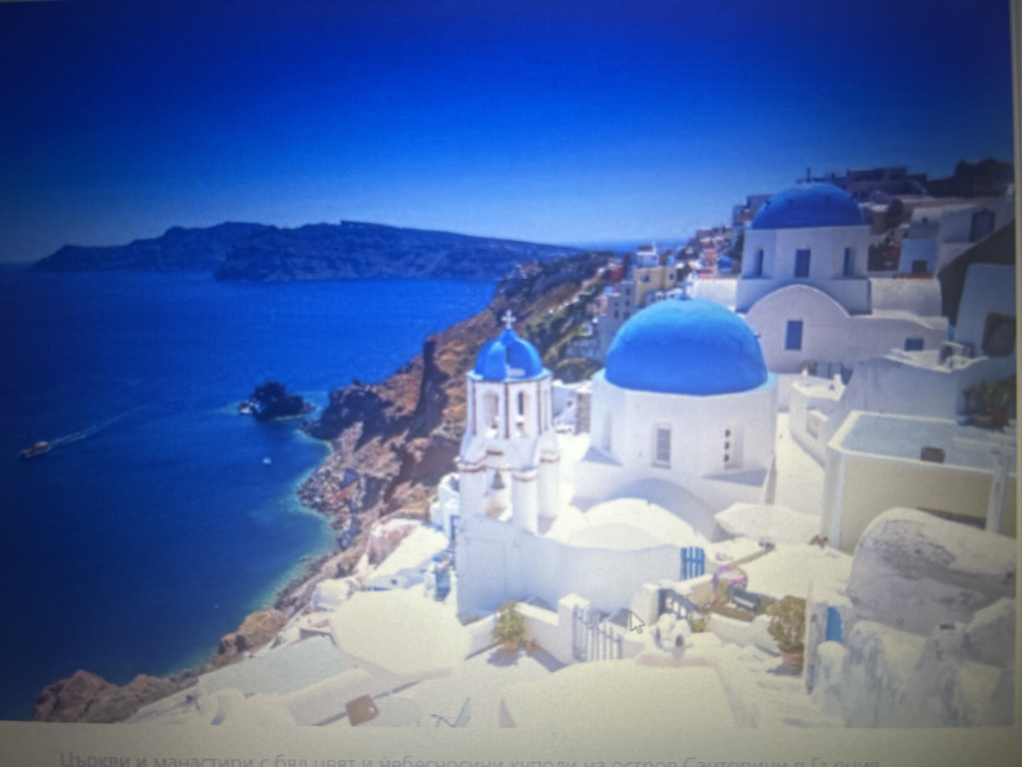 The image depicts a scenic view of a white village with blue-domed churches overlooking a deep blue sea. The architecture is characteristic of the Greek islands, with buildings clinging to a cliffside. The sky is a vibrant, cloudless blue. In the distance, a rugged coastline and a few small boats are visible on the water. The foreground shows pathways and small potted plants, adding to the picturesque setting. The provided image and hints do not contain any information or visual cues that would place this scene within the city of Sofia, Bulgaria.