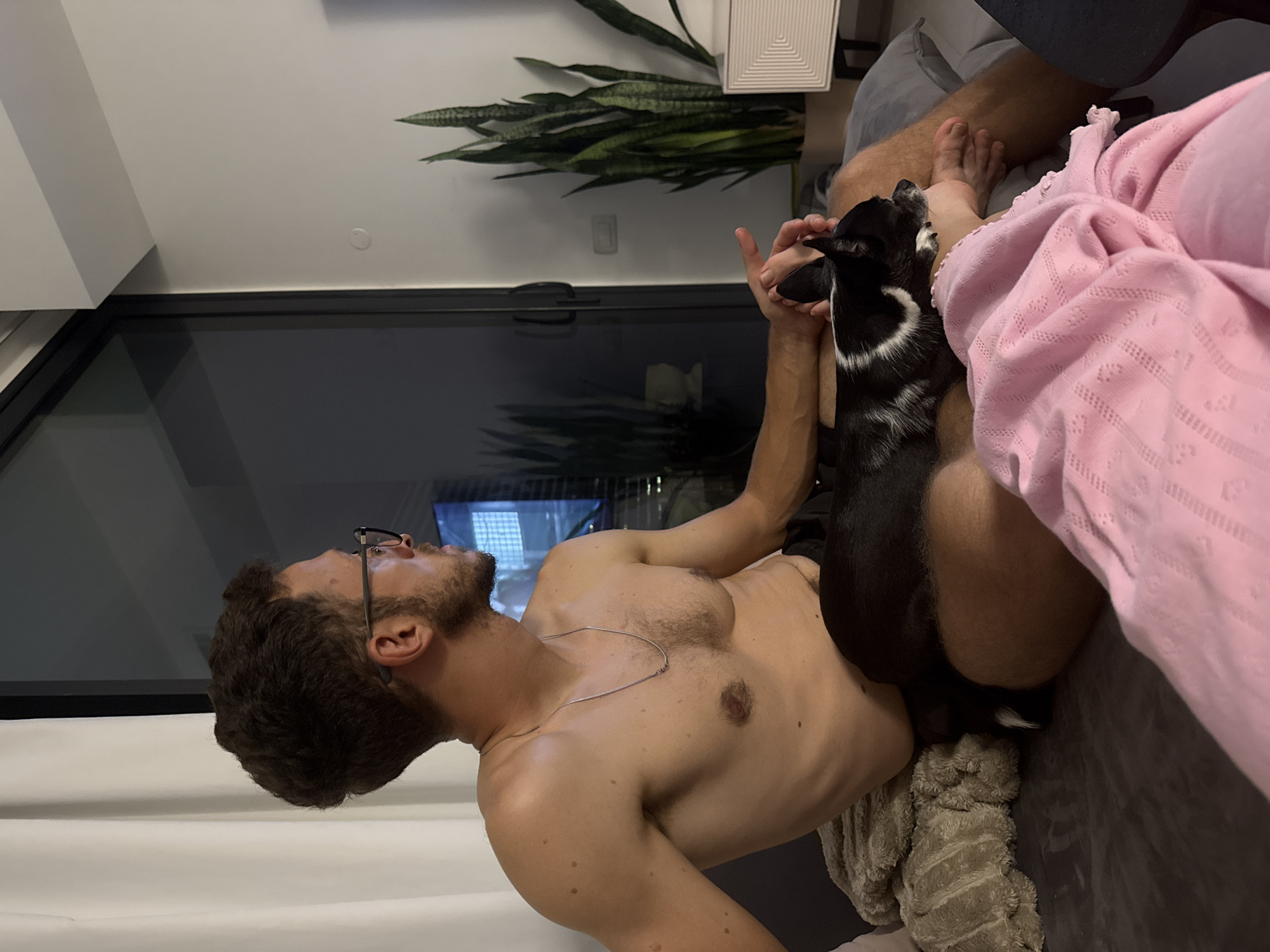 A shirtless man with glasses is lying down indoors. A small black and white dog stands on him, possibly interacting with his hand. Part of another person is visible, covered by a pink fabric. A snake plant sits on a white surface in the background. The scene is indoors, possibly in an apartment, and the dark windows suggest it could be night or a dimly lit space. There is no information in the image to indicate this picture was taken in Ramat Gan, Israel.