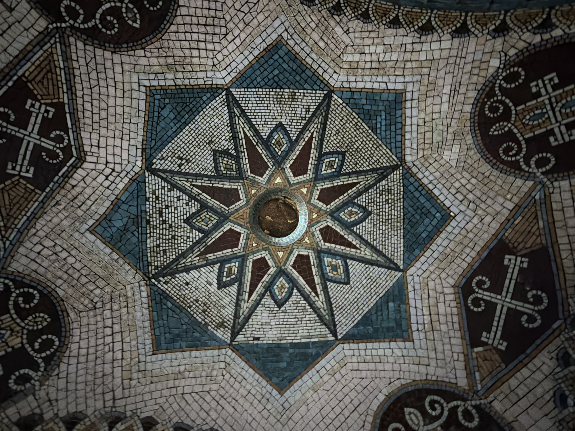 This image shows a mosaic floor in a building in Tbilisi, Georgia. The mosaic features a star pattern at its center, surrounded by geometric shapes and decorative symbols, all composed of small, square tiles in varying shades of blue, brown, and white. The overall design suggests a historical or religious significance, possibly located in an old building or public space.