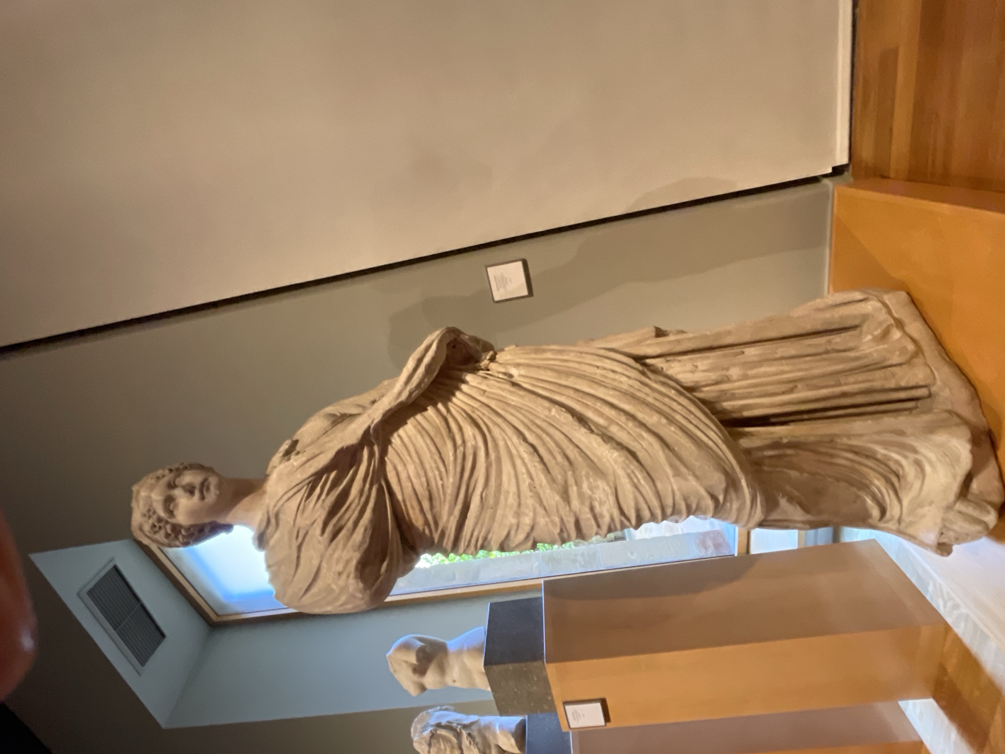 A stone sculpture of a draped female figure stands on a wooden plinth in what appears to be a museum or gallery setting. The sculpture's location in Barcelona, Spain, cannot be verified from the image. The room has a light grey wall, a white ceiling, and a wooden floor. The artwork is lit from above, possibly by a skylight or window, highlighting the folds of the drapery and the figure's face. The sculpture stands above at least two other sculptures. The area in front of the sculpture is brightly lit, whereas the area surrounding the plinth is in shadow. A rectangular placard hangs on the wall behind the sculpture.