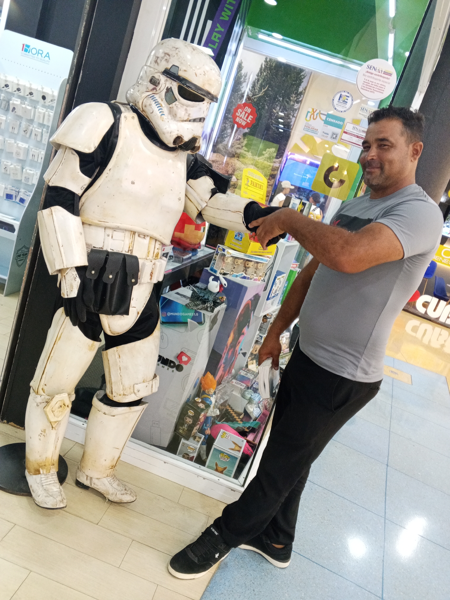 A man in a gray t-shirt and black pants stands indoors, looking at a person in a weathered white stormtrooper costume. The stormtrooper, with a dark undersuit, holds a black object in its gloved hands, extended towards the man. The man's right arm is extended as well, appearing to be interacting with the stormtrooper. The background suggests a retail environment, with shelves of products visible behind a glass display case. The man's left foot is slightly behind his right, and he is looking towards the stormtrooper. The stormtrooper is facing the man, with its head tilted slightly. The scene is brightly lit, possibly in a mall or a store.