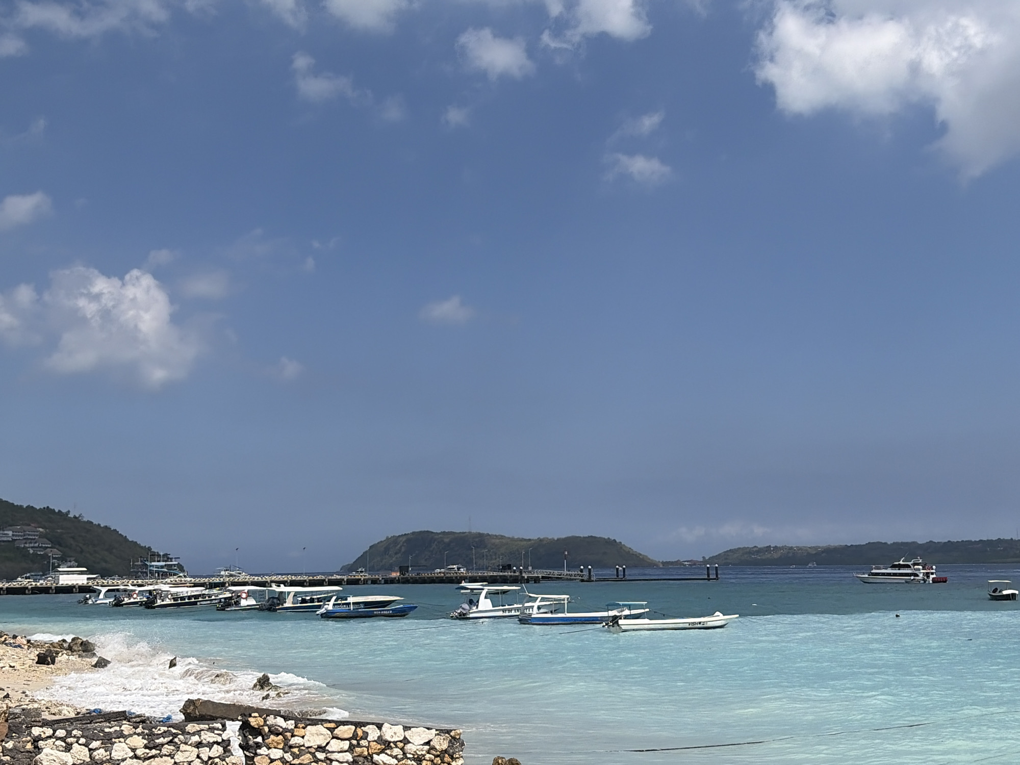 A wide shot captures a bright, sunny day at a coastal area, likely in Nusa Penida, Indonesia, with clear blue skies dotted with fluffy white clouds. The turquoise waters are calm, with small waves lapping at a rocky and sandy shore in the foreground. A low, dark stone wall is visible along the immediate edge of the beach.
A long pier extends into the water from the left side of the image, lined with numerous boats. These include several speedboats with white hulls and blue accents, some appearing to be passenger ferries, docked side-by-side. Further out in the water, more boats are anchored or moving. In the distance, a larger white passenger ferry is visible, along with a small white boat on the far right.
The background features green, hilly islands with scattered buildings on the slopes, adding to the tropical ambiance. The overall scene depicts a bustling harbor or ferry terminal in a scenic coastal location.