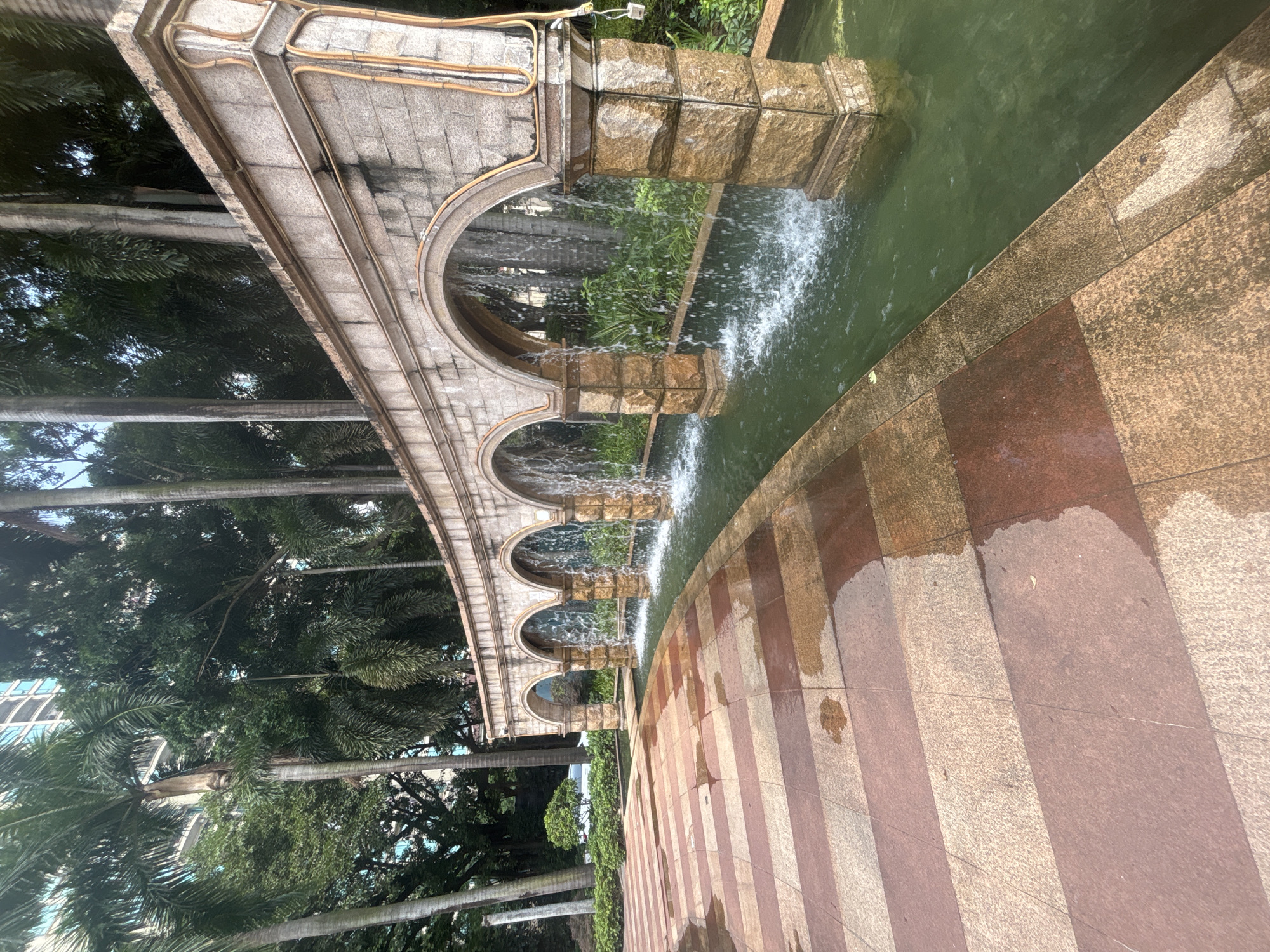 The image shows an outdoor scene with a decorative stone fountain structure. The fountain features a series of arches, through which water cascades into a shallow green pool. The structure is made of light-colored stone blocks with darker, reddish-brown accents. Behind the fountain, lush green foliage, including palm trees, is visible, with a tall building in the background. The foreground consists of a tiled pathway with a repeating pattern of alternating reddish-brown and light-colored rectangular tiles. The tiles appear wet, suggesting recent rain or the fountain's spray. No people are present in the image. The setting appears to be a well-maintained public space within a city, likely in a tropical or subtropical region given the presence of palm trees. This location is Nanhai District, China.