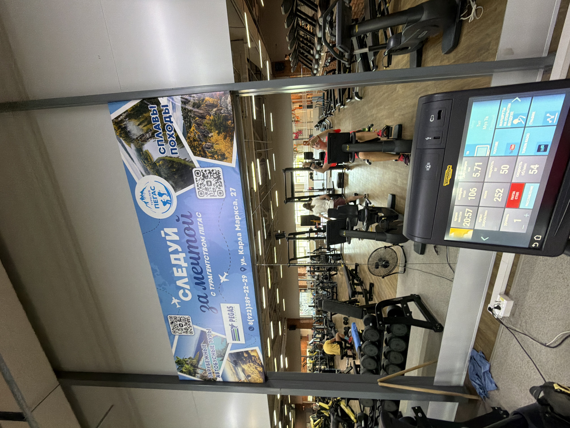 The image depicts the interior of a gym, specifically a Technogym branded stationary bicycle. The console of the bicycle displays data such as time, calories, distance, and speed. In the background, other gym equipment, including treadmills and weight machines, is visible. Several individuals are engaged in workouts, with one person in the foreground taking a selfie in the mirror. A large advertising banner is prominently displayed on the wall, featuring images of natural landscapes and travel-related text in Russian. The banner includes the text "СЛЕДУЙ за мечтой" (Follow your dream), "С ТУРАГЕНТСТВОМ ПЕГАС" (with travel agency Pegasus), and contact information including a phone number and an address: "8(922)389-22-29 ул. Карла Маркса, 27" (8(922)389-22-29 Karl Marx Street, 27). This address and phone number strongly suggest the location is in Chaikovsky, Russia. The overall environment appears clean and well-lit, characteristic of a modern fitness facility.