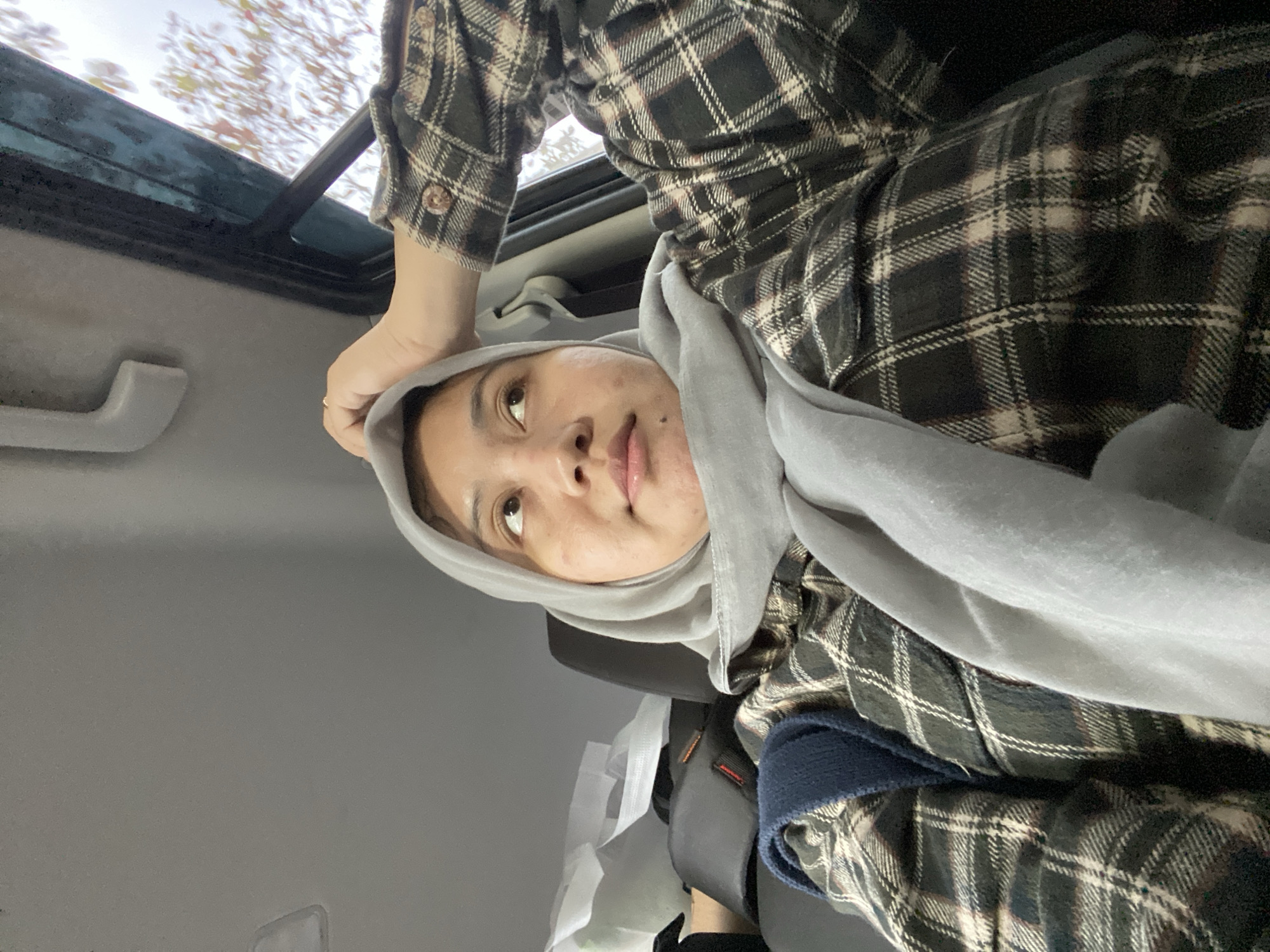 A person wearing a gray hijab and a plaid shirt is sitting in the passenger seat of a vehicle, looking upwards and to the side. Their right hand is resting on the top of their head. The interior of the car is visible, including a grab handle and part of the window, which shows a blurred background of trees. Bags are piled up in the back seat. The scene appears to be captured outdoors, possibly in the city of Banjarbaru, Indonesia.