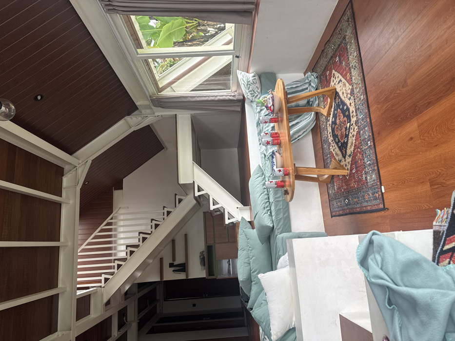 The image shows a multi-level interior space featuring wooden and metal structures. A white staircase leads to upper levels. A skylight provides natural light. The interior includes a couch with light blue cushions and a wooden coffee table holding multiple cans. A patterned rug lies on the wooden floor. The perspective is a top-down view, and the image is rotated.